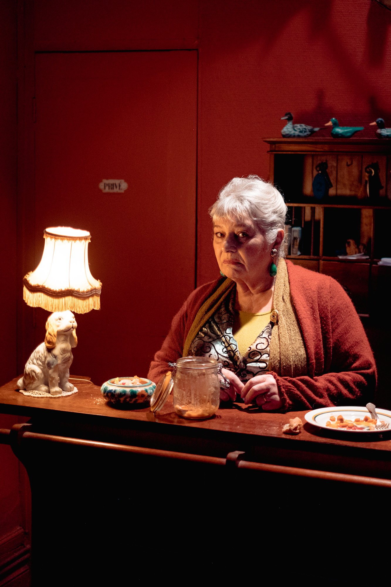 Une vieille femme assise derrière un comptoir en bois, éclairée par une lampe de bureau en forme de chien, dans une pièce avec des décorations en bois et des objets divers en arrière-plan.