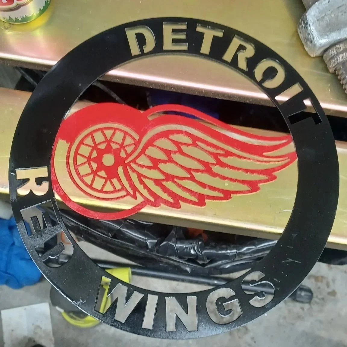 A black metal circular sign with cut-out text reading 'Detroit Wings' and a stylized red Detroit Red Wings logo in the center, which features a winged wheel.