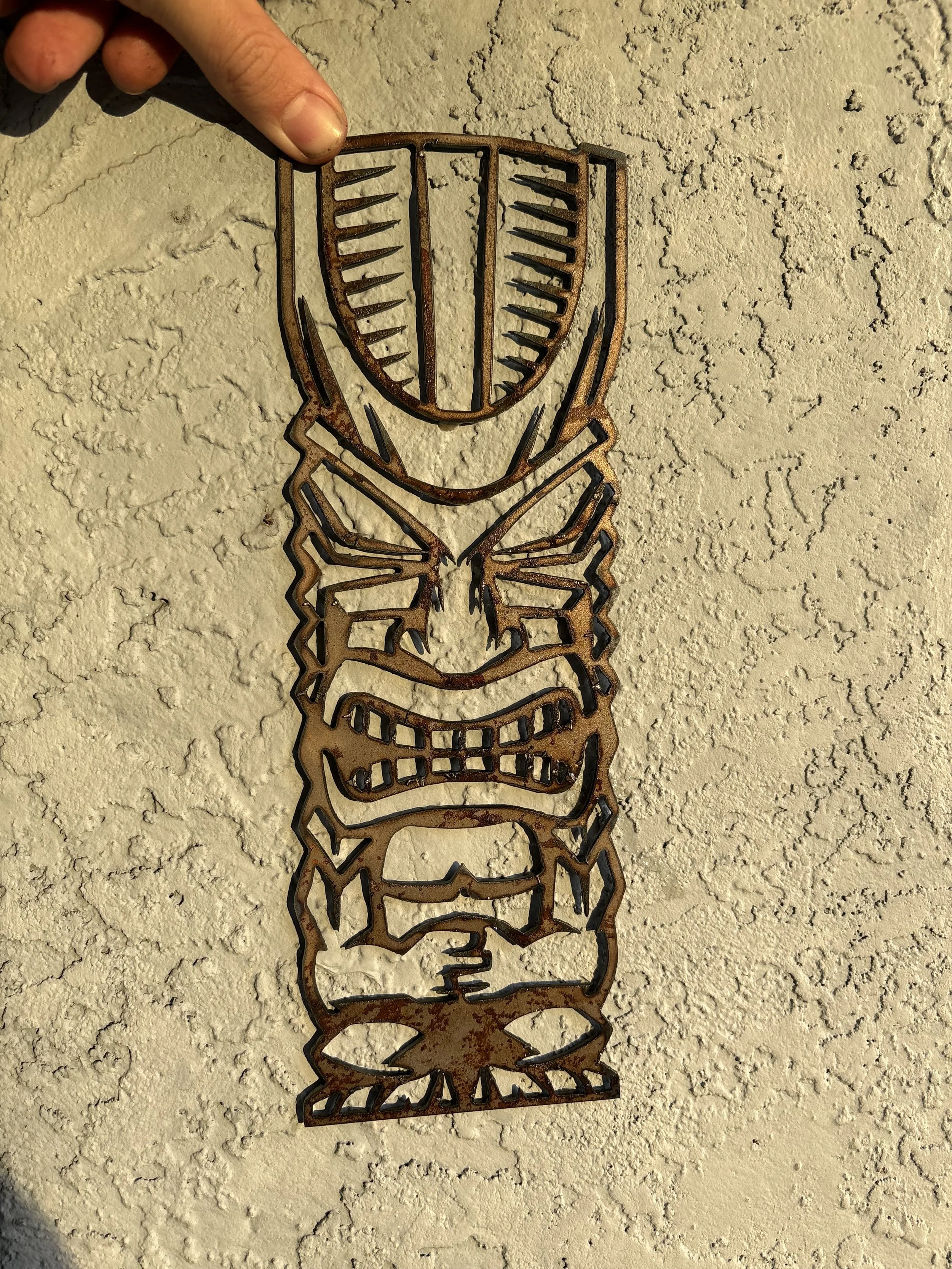 A metal wall art sculpture of a stylized face or mask, held by a person's hand, against a textured beige wall.