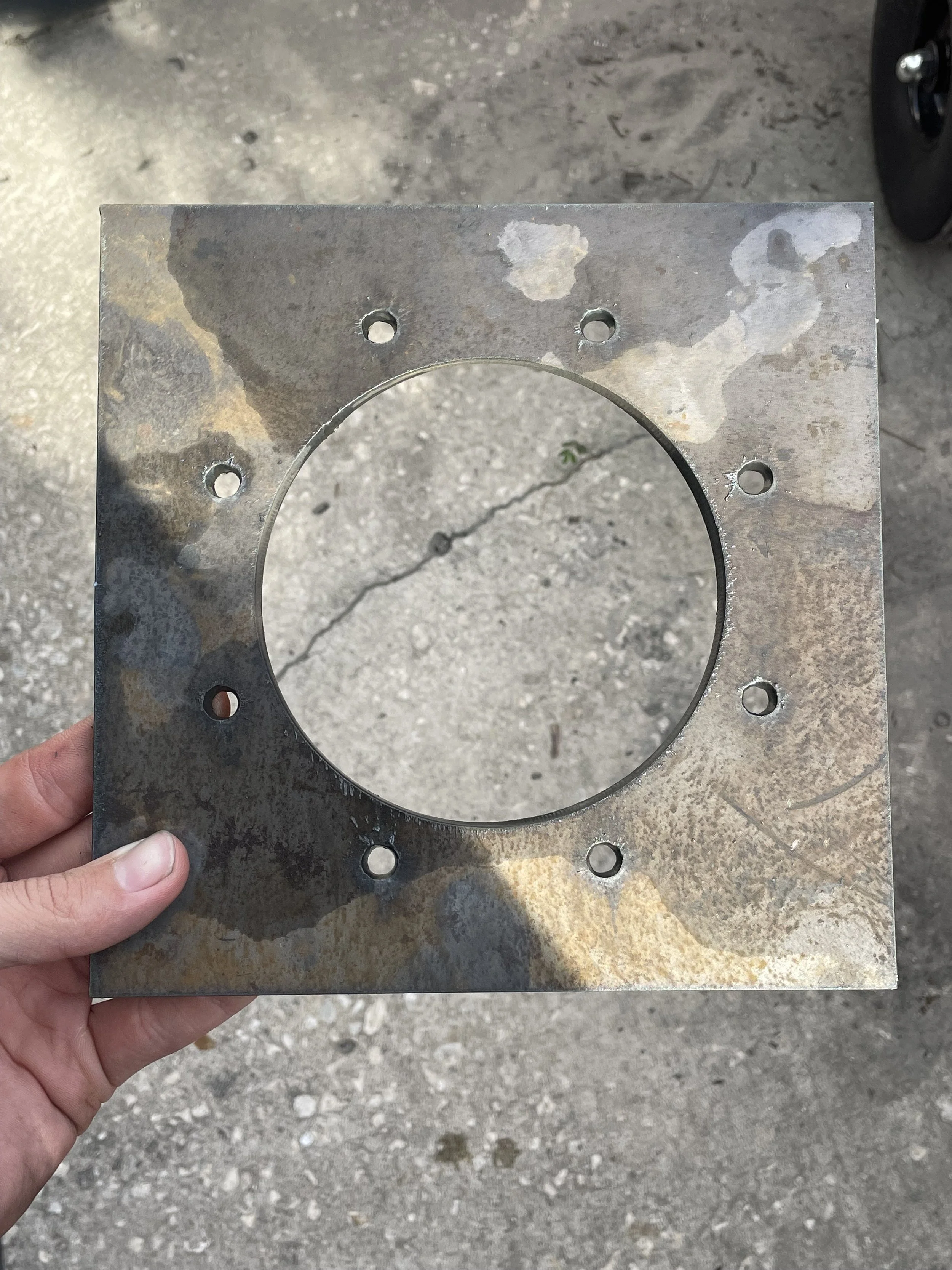 A person holding a square metal plate with a large circular hole in the center and multiple smaller holes around it, outdoors on a concrete surface.