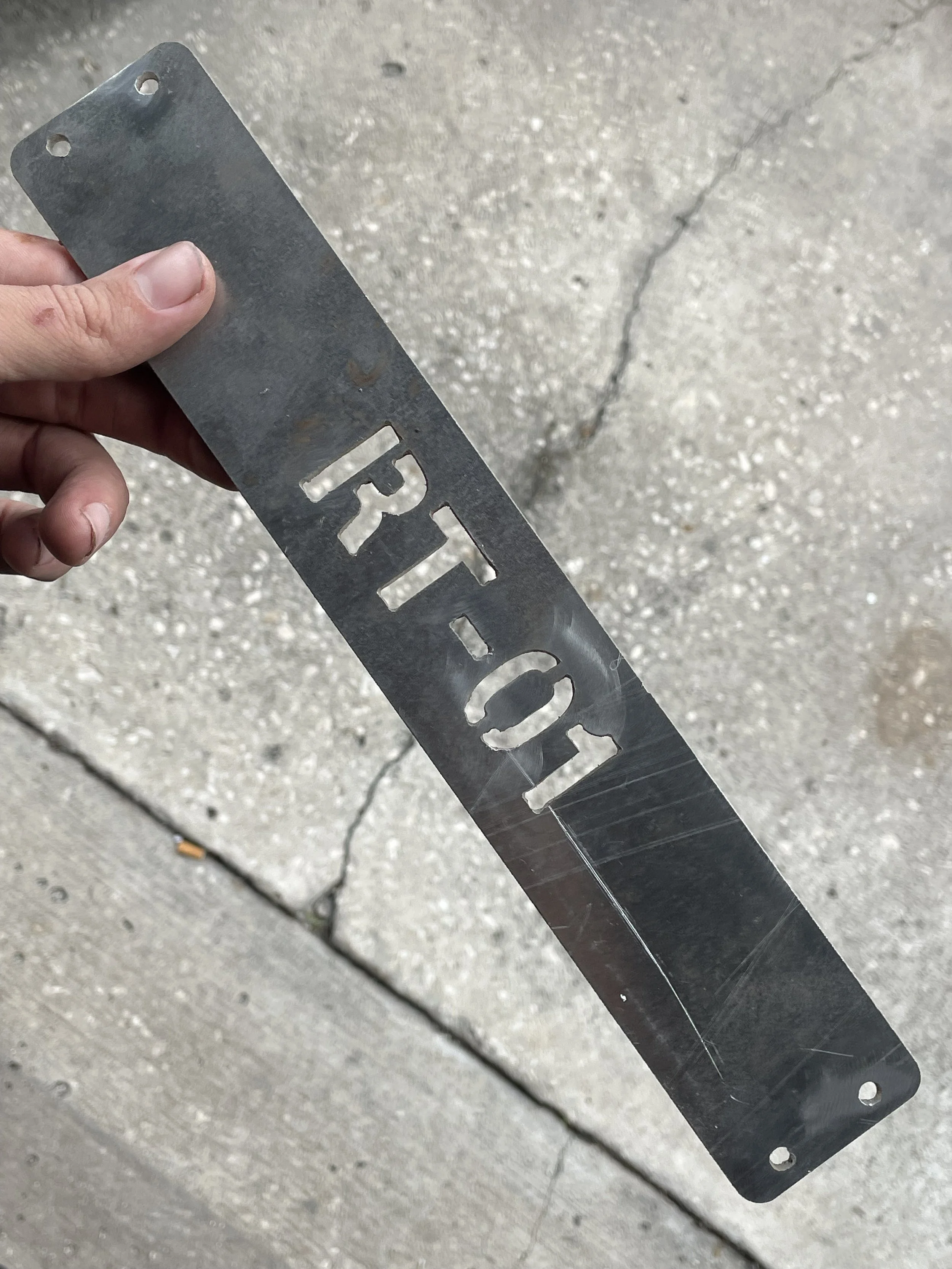 A person holding a metal license plate frame with holes for mounting and the words 'RT-01' cut out, above a concrete sidewalk.