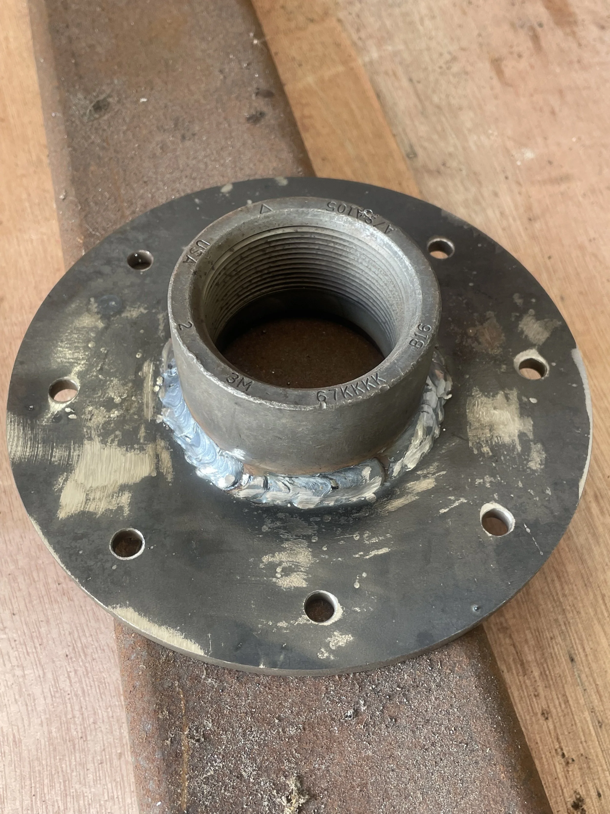 Metal component with a central threaded hole attached to a circular base with multiple bolt holes, placed on a wooden surface.