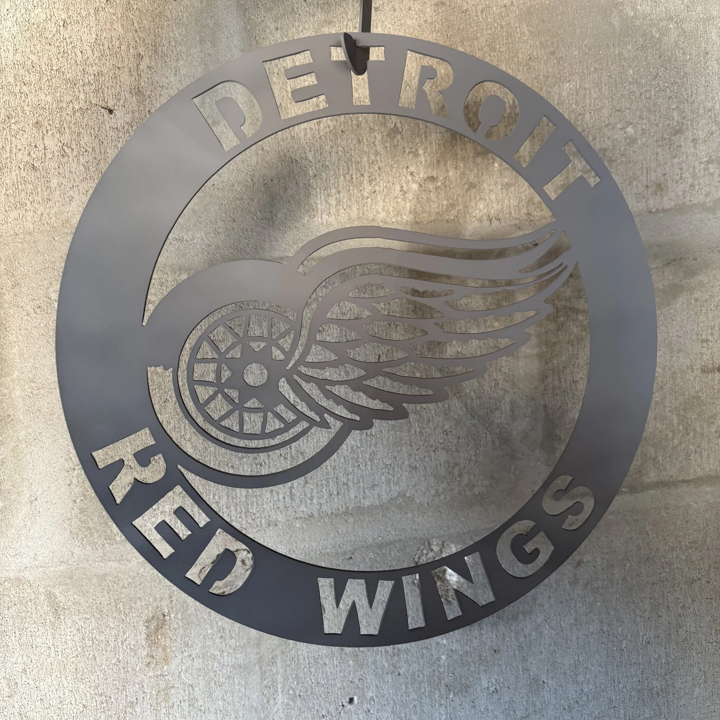 Metal Detroit Red Wings logo with winged wheel design on a concrete wall.