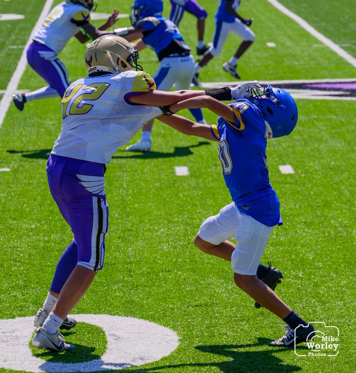 Two young football players in blue and white uniforms are engaged in a physical tackle during a game on a green football field, with other players visible in the background.