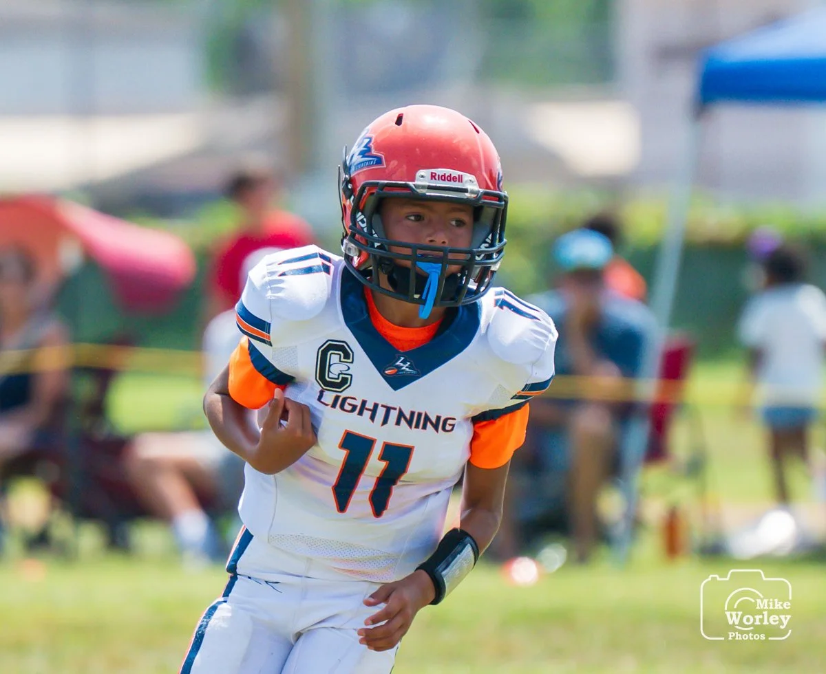 Young football player in white and orange uniform running with a football, wearing a red helmet with a faceguard, during a game on a grassy field with spectators in the background.
