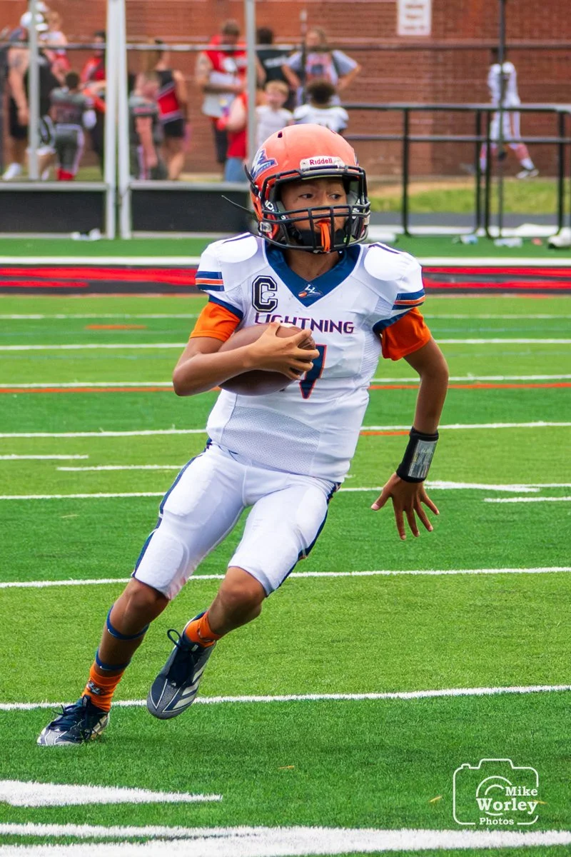 A young football player in a white and blue uniform running with the ball on a football field. The player is wearing a helmet and has a wrist band on their right arm. There are spectators in the background.