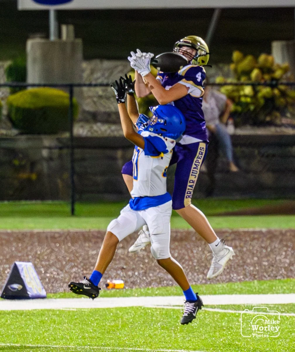 Two young football players, one in purple and one in white and blue, compete for possession of the football in a night game. The player in purple reaches for the ball while the player in white and blue jumps to catch it.