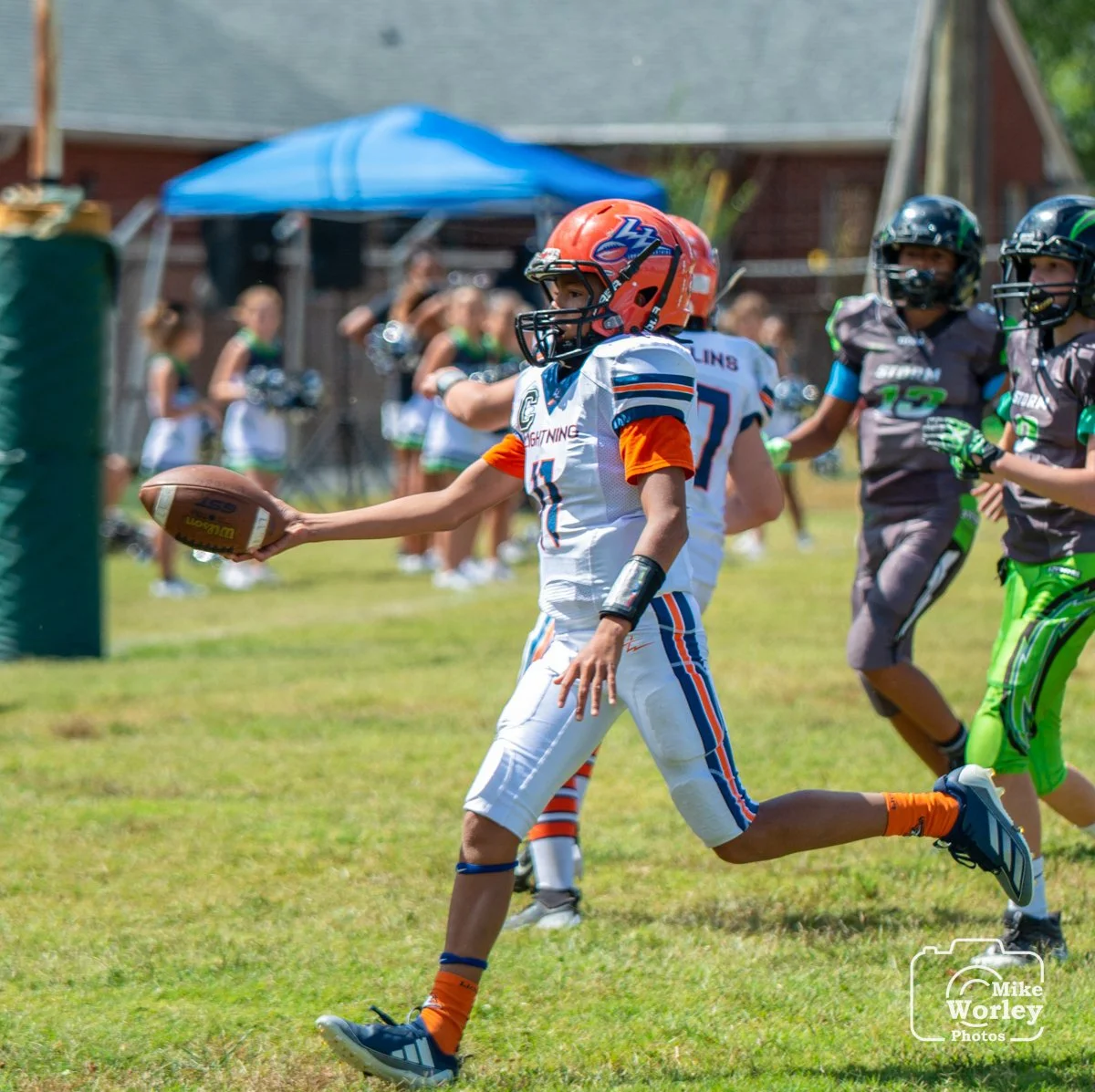 A young football player in a white and orange uniform runs on a grassy field holding a football, with other players and cheerleaders in the background.