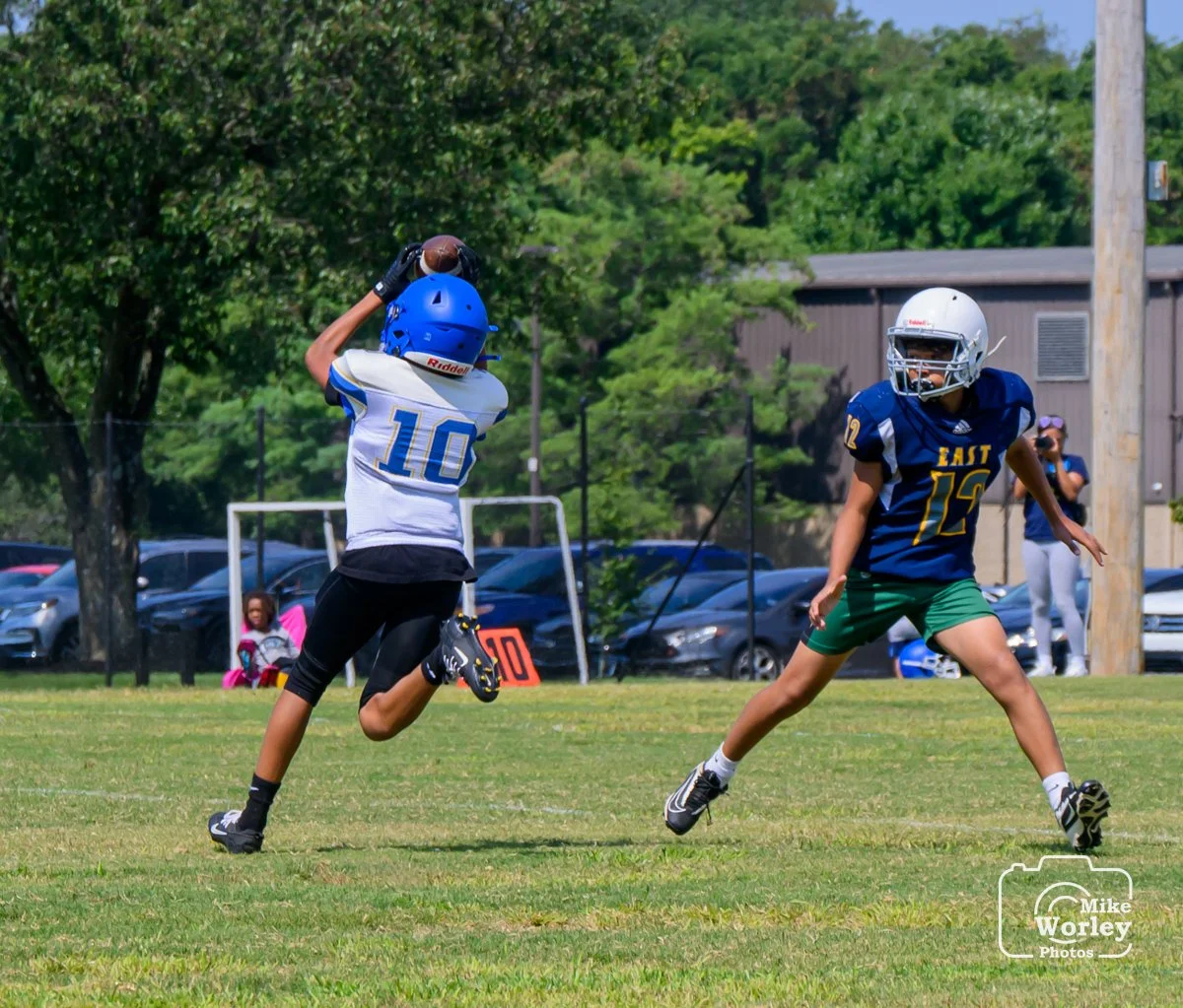 A youth football player catching a pass while another player on defense approaches on a grassy field during a game.