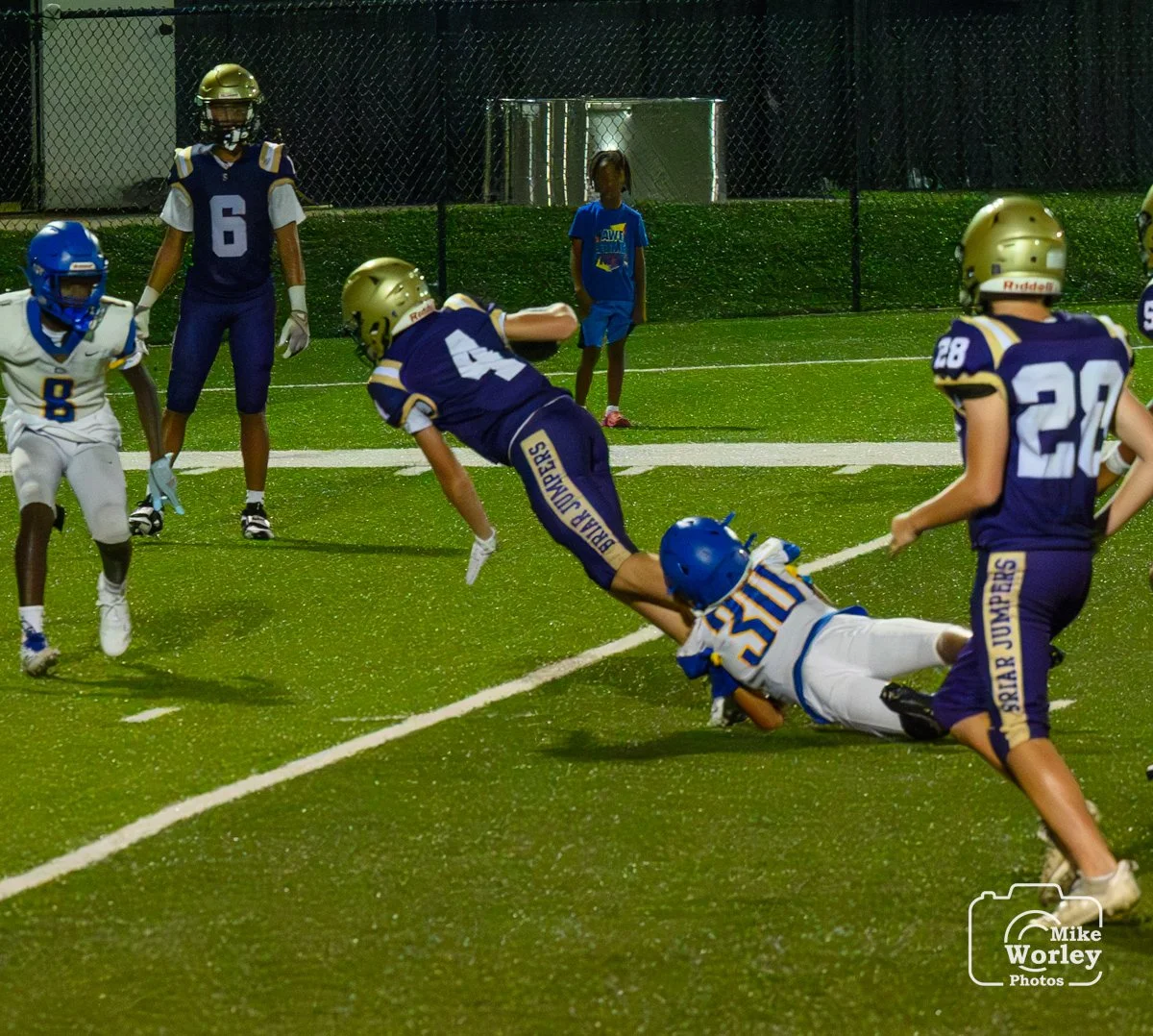 Youth football game with players in blue and white uniforms on a green field, one player being tackled by a blue and white player, officials and spectators in the background.