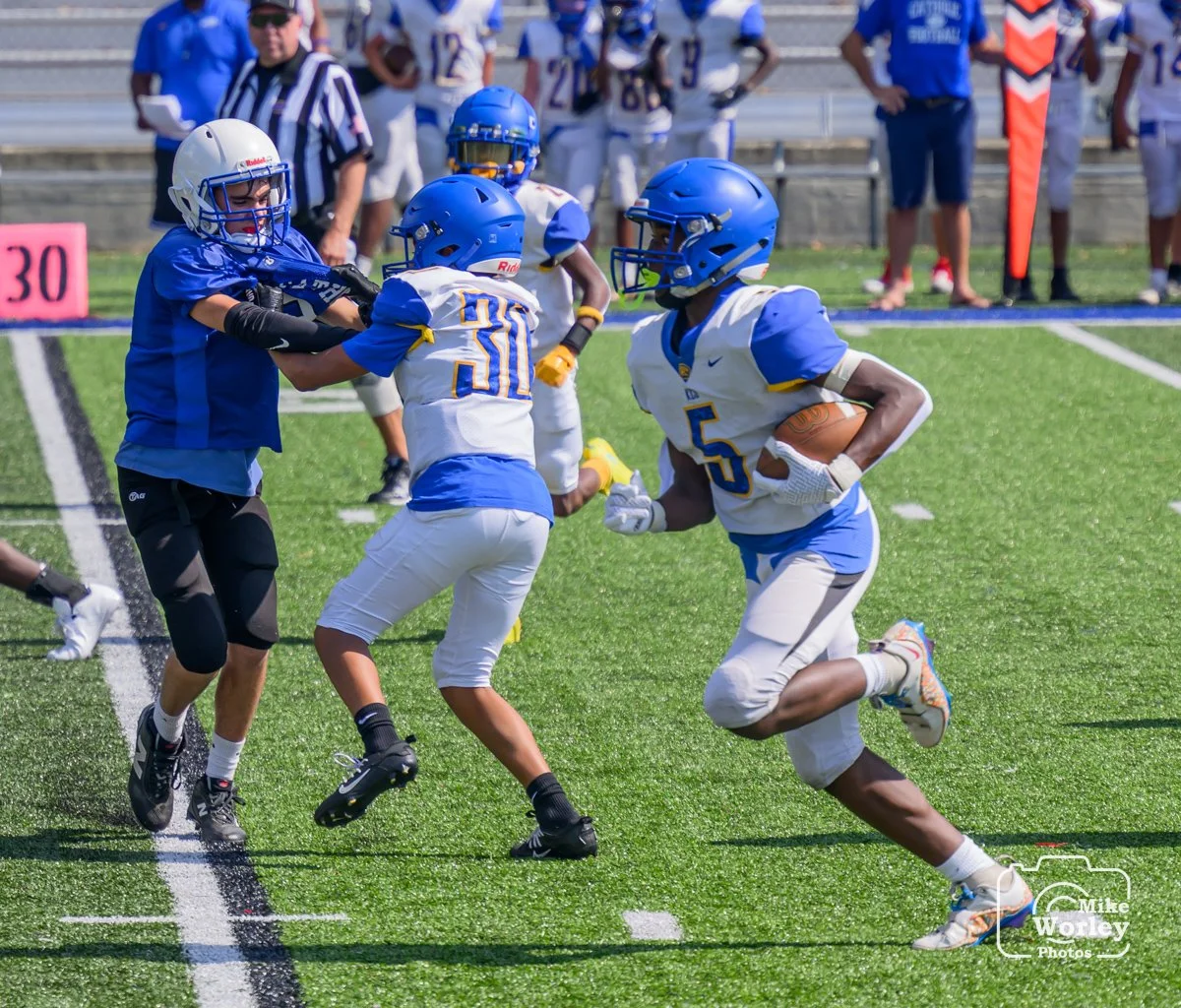 A youth football game in progress with a player running with the ball while defenders try to stop him, on a green turf field with sideline and officials watching.