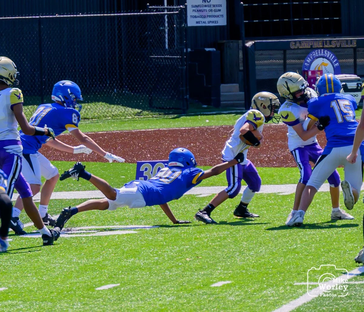 Youth football game with players in blue and yellow jerseys, one player diving to tackle another with the football, on an outdoor field.