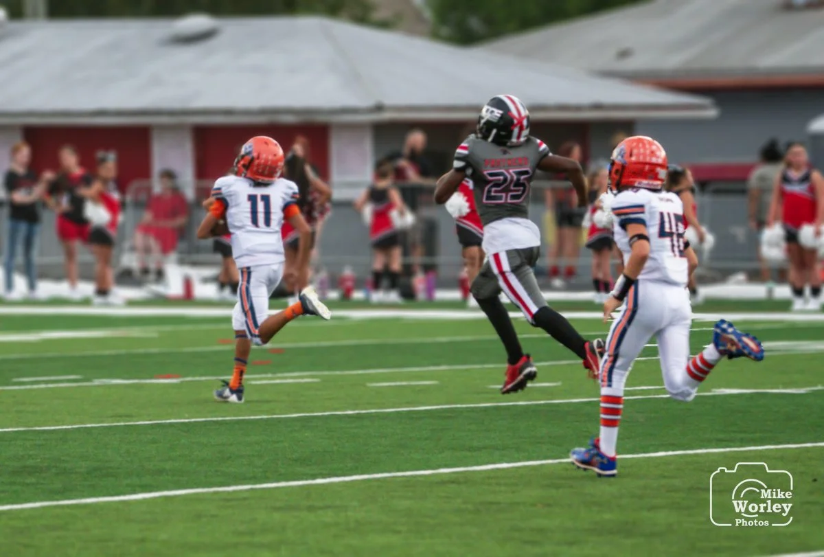 Children playing youth football on a field, wearing helmets and jerseys, with spectators watching in the background.