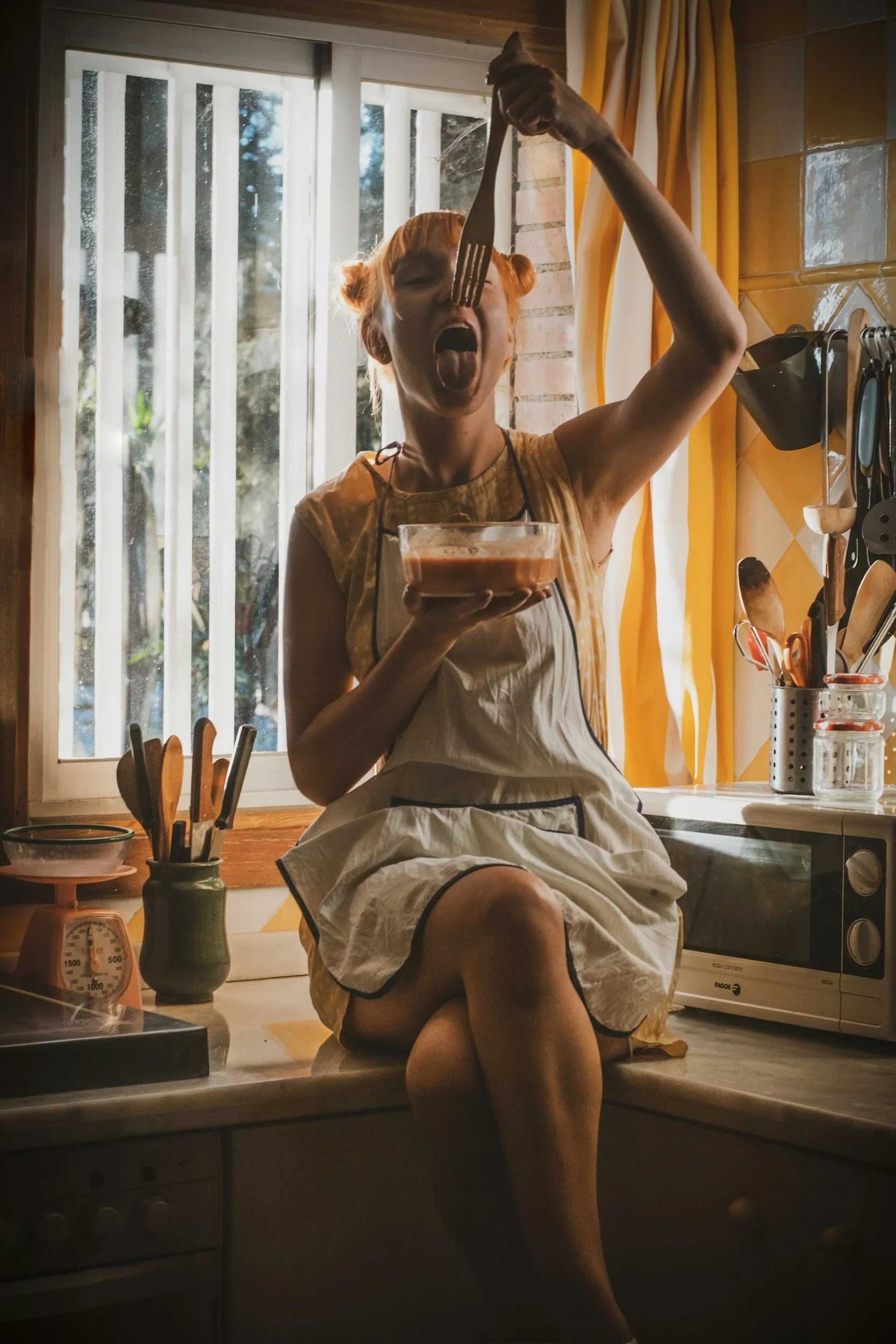 A woman with red hair styled in buns, wearing an apron, sitting on a kitchen counter and holding a bowl of food in one hand while striking a playful pose with a fork in her mouth, in a cozy kitchen with sunlight coming through a window.