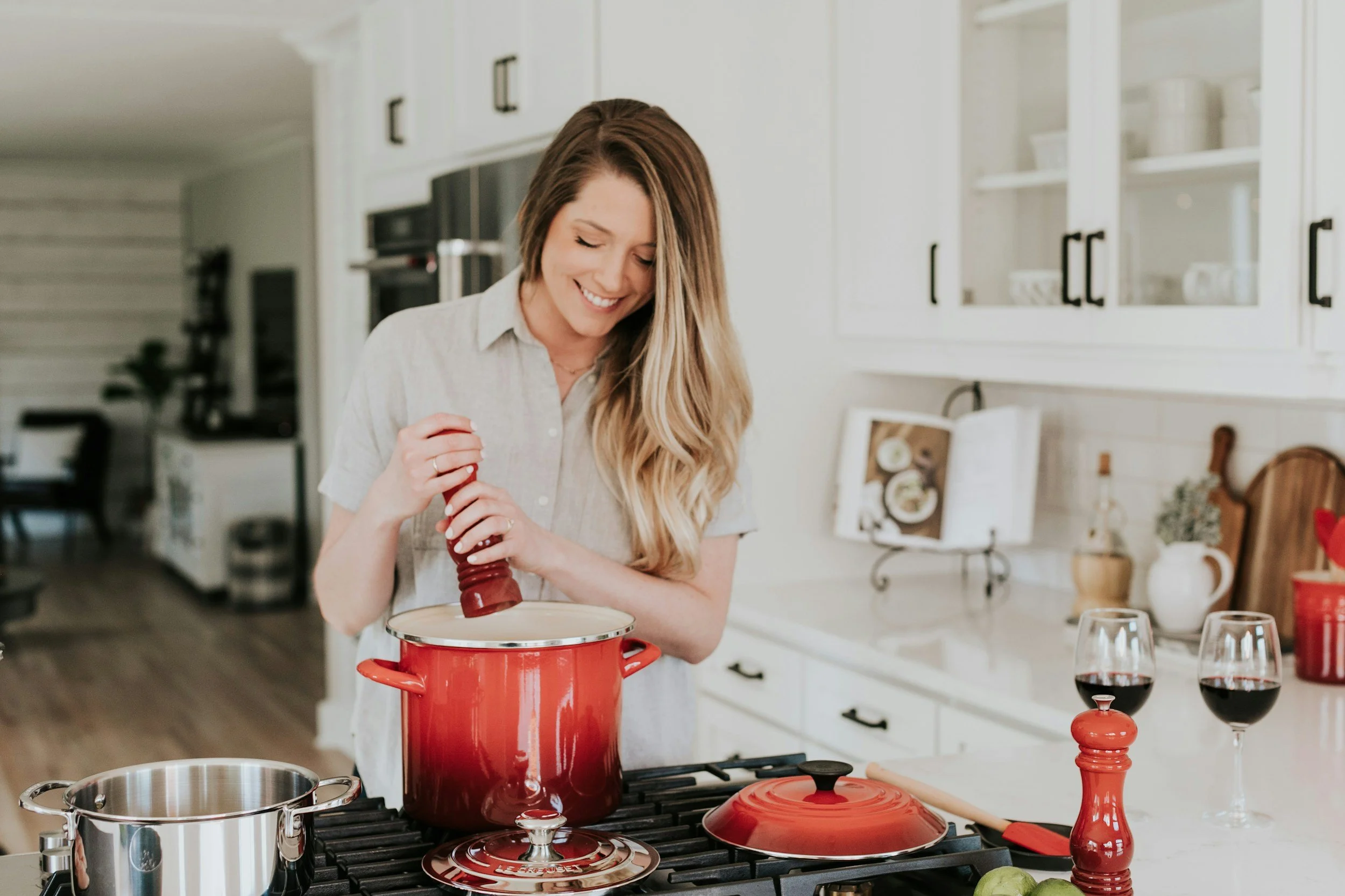 A woman smiling and cooking in a bright kitchen with a red pot on the stove, holding a pepper grinder, with two glasses of red wine on the counter.