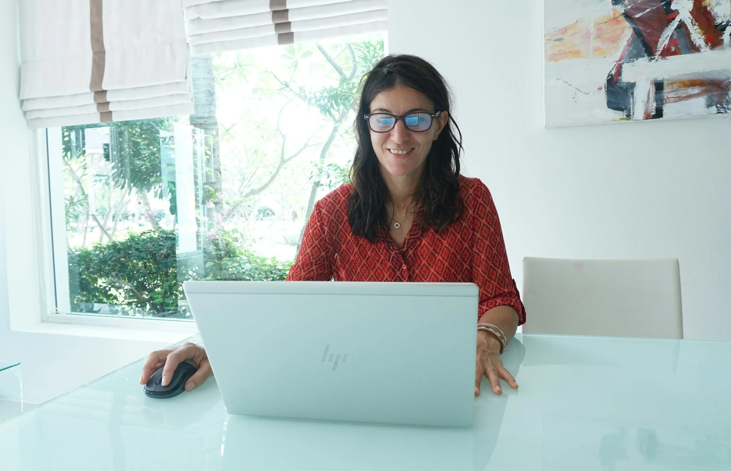 Woman with dark hair, glasses, wearing a red patterned shirt, sitting at a glass table working on a silver HP laptop, with a mouse in her right hand, in a bright room with a window and abstract art on the wall.