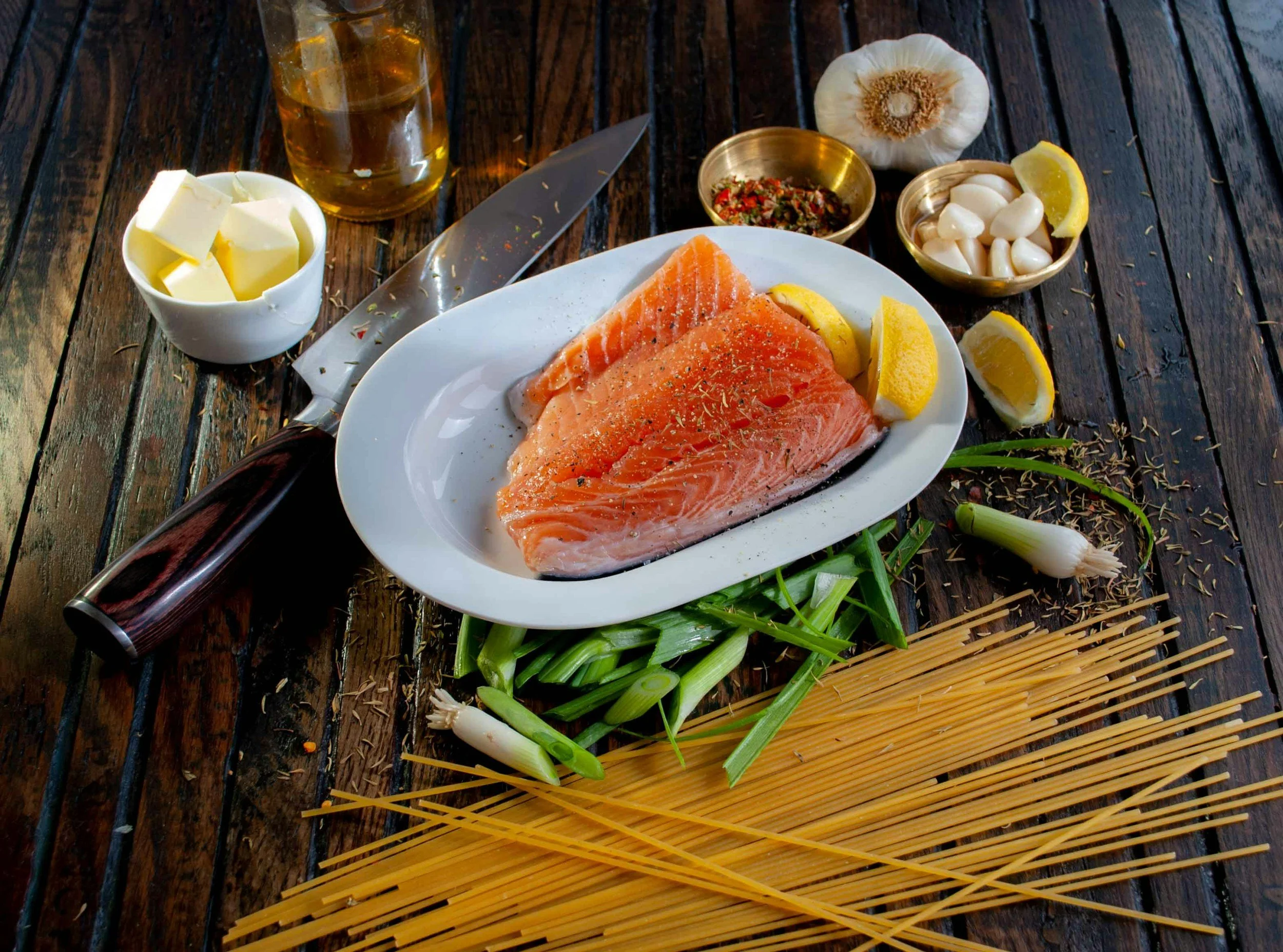 Raw salmon fillets on a white plate with lemon wedges, surrounded by green onions, garlic, herbs, spaghetti pasta, butter, and seasonings on a wooden table.