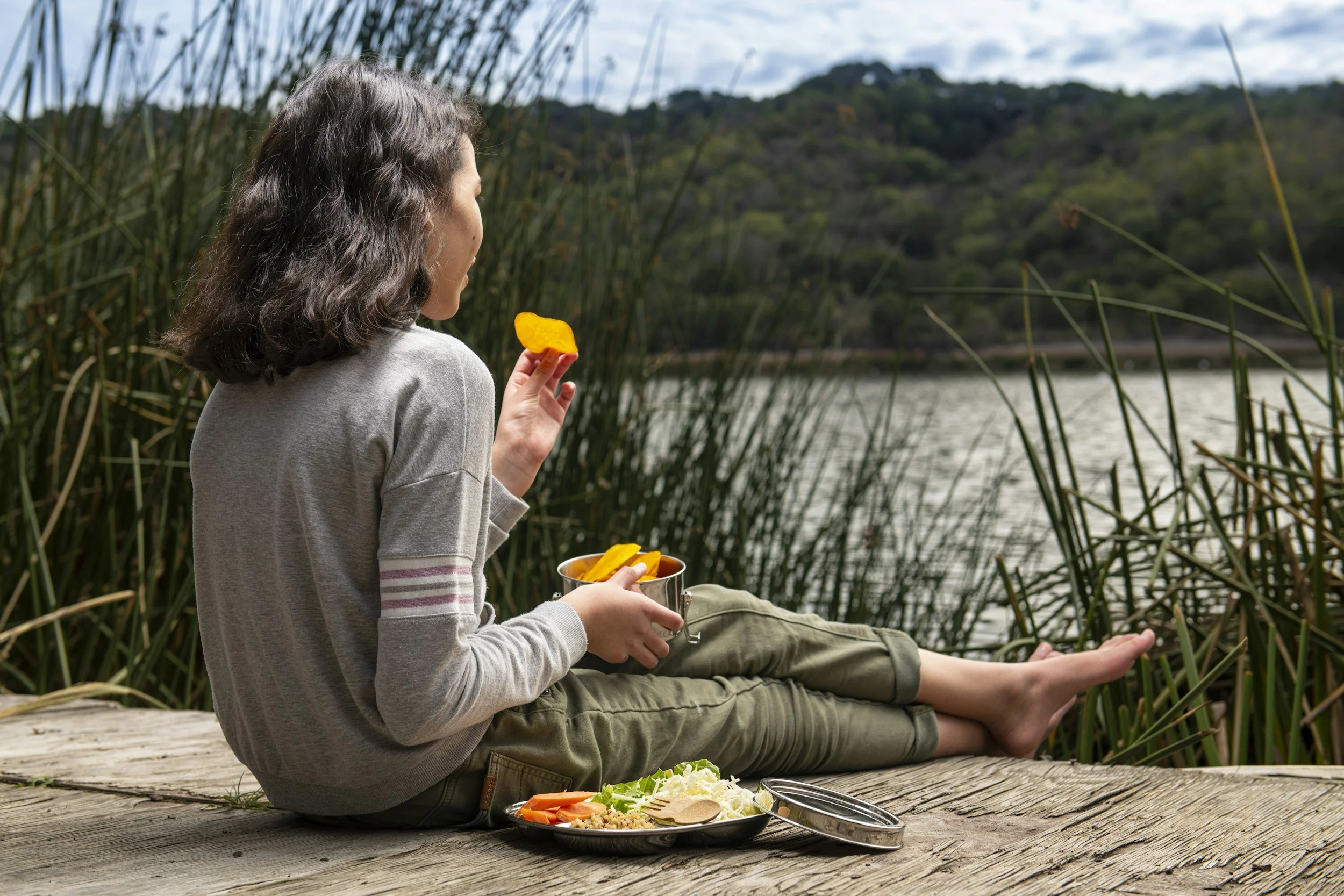 A woman sitting on a wooden dock by a lake, holding a bag of sliced mango and eating one. She has a plate of salad, vegetables, and rice in front of her, with more mango slices in a bowl nearby. The scene is surrounded by tall grass and trees with a cloudy sky overhead.