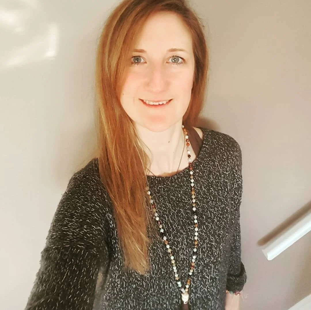 A woman with fair skin and red hair smiling at the camera, wearing a black sweater with white specks and a beaded necklace, standing against a beige wall.