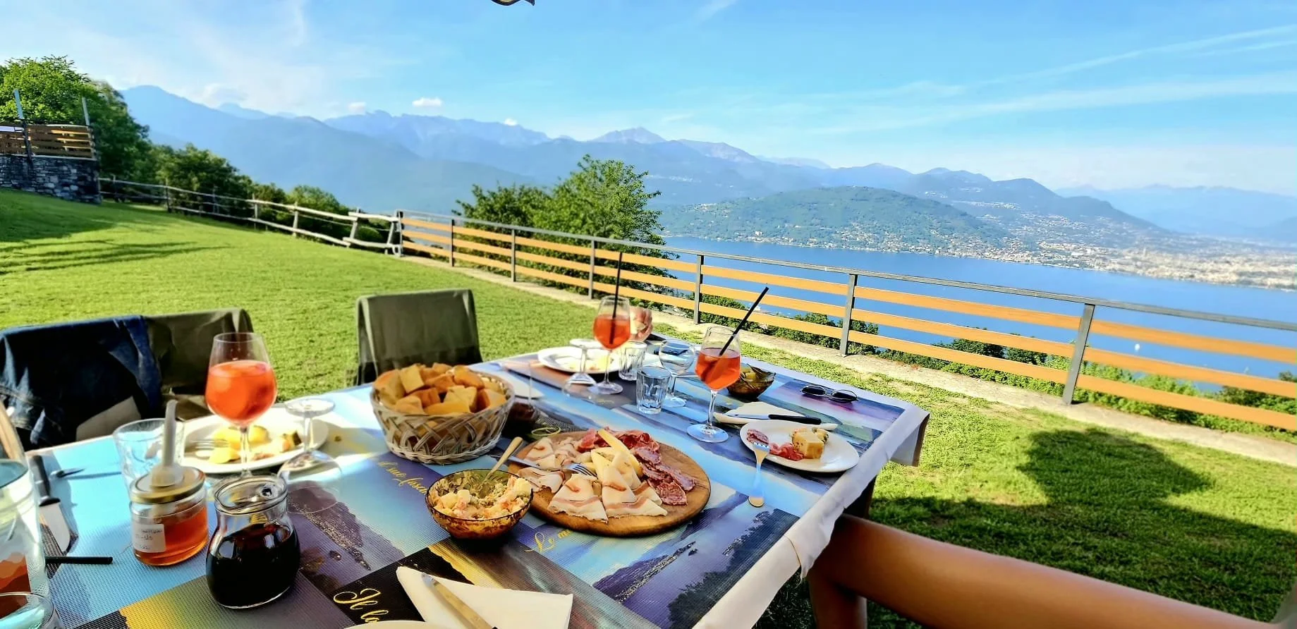 Tavola apparecchiata con cibo e drink davanti a un panorama di monti, lago e cielo azzurro con nuvole, in un giardino con prato verde e recinzione.