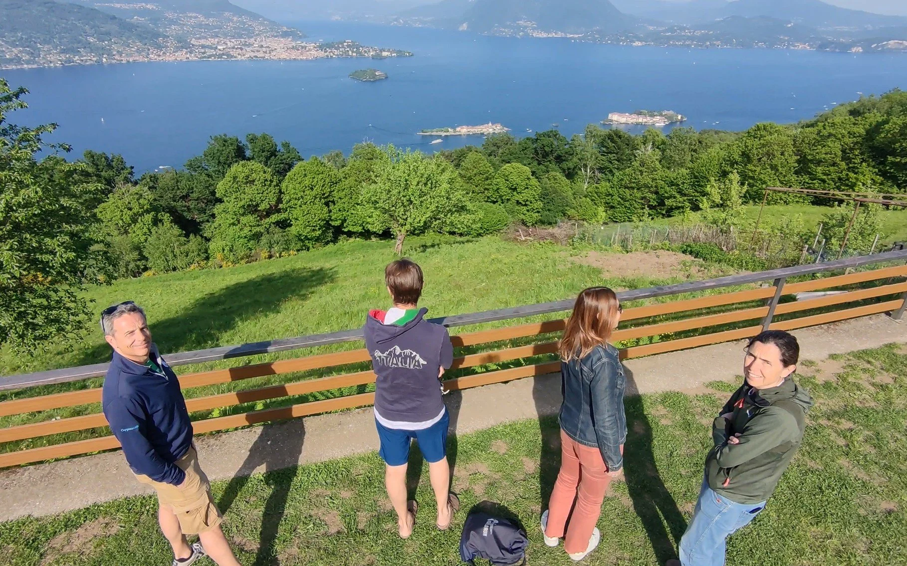 Cinque persone si trovano vicino a una ringhiera di legno in un'area verde con vista su un grande lago e montagne, alcune persone sono rivolte verso il lago e altre si ribaltano.