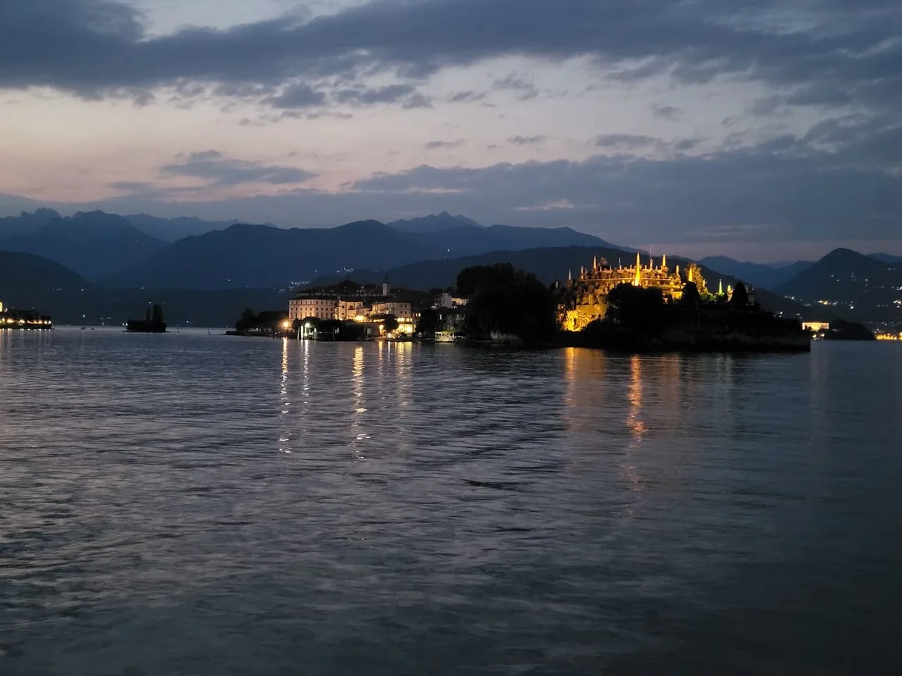 Veduta notturna di un lago con un'isola illuminata, montagne sullo sfondo e un cielo nuvoloso.