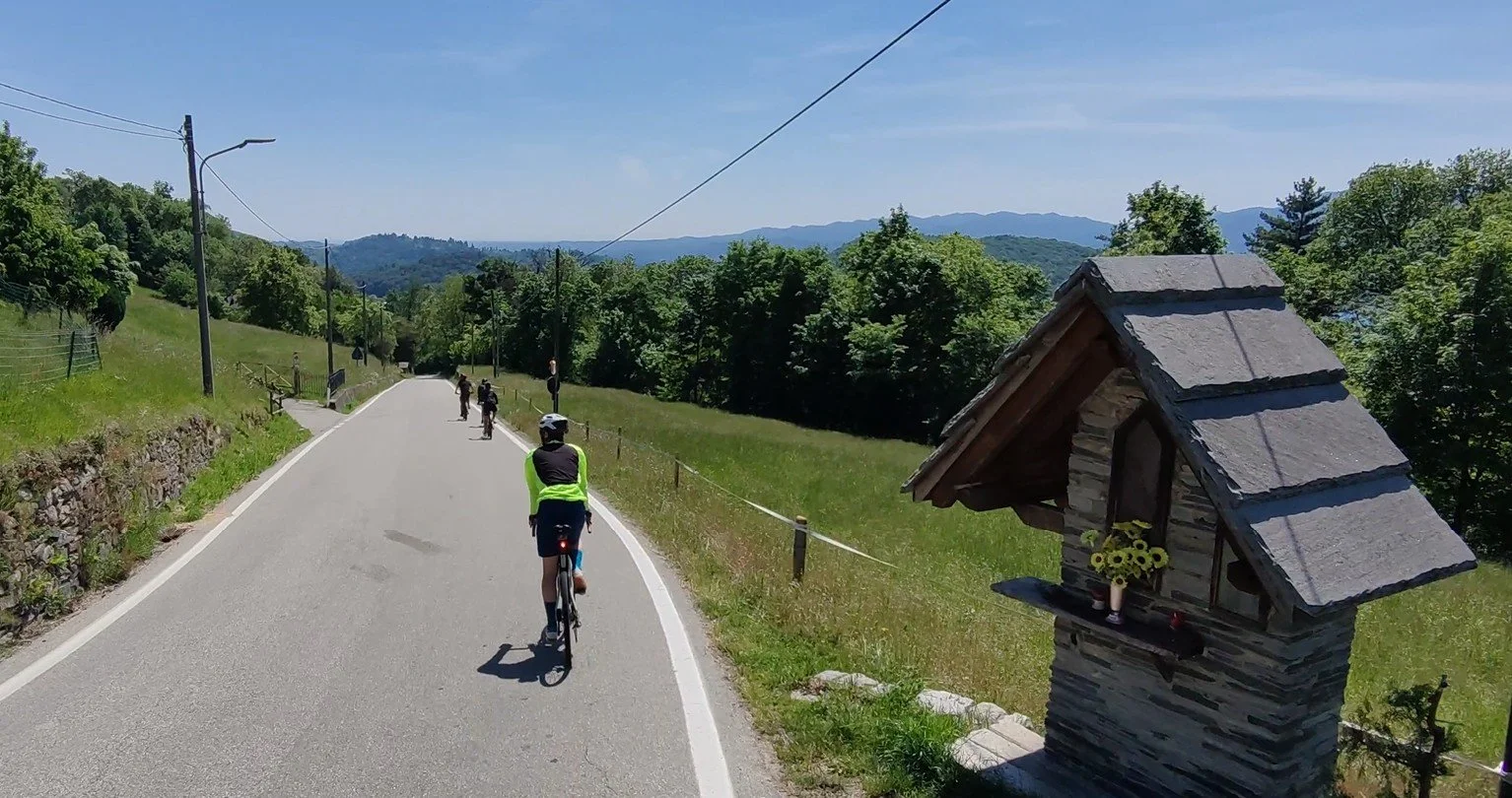 Strada di campagna con ciclisti che pedalano, paesaggio verde e montagne sullo sfondo, piccolo edificio in pietra con tetto di tegole, sicuro e soleggiato