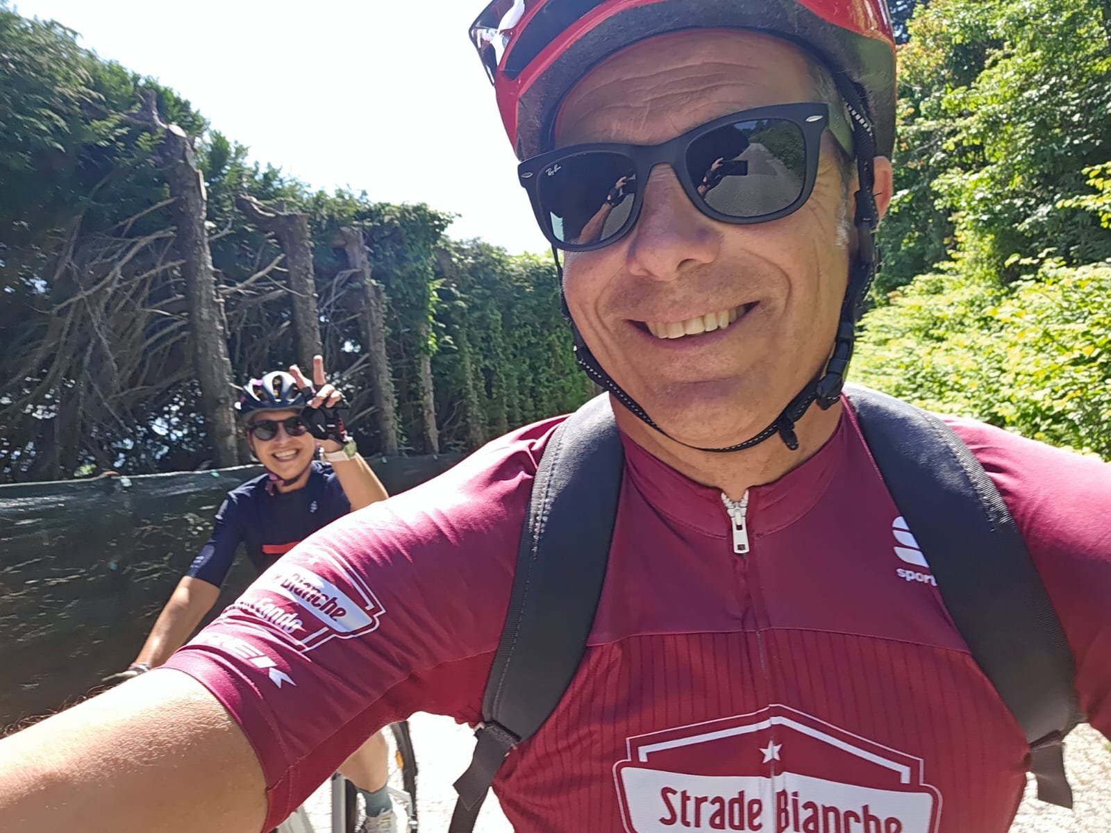 Due ciclisti sorridenti avanzano su un sentiero immerso nella natura con alberi e foliage verdi. La persona in primo piano indossa occhiali da sole, casco e maglietta rossa, mentre la seconda ciclista saluta la fotocamera e indossa occhiali da sole, 