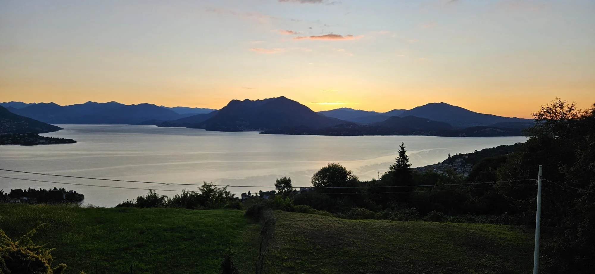 Paesaggio naturale al tramonto con un lago, montagne sullo sfondo e alberi e vegetazione sul primo piano.