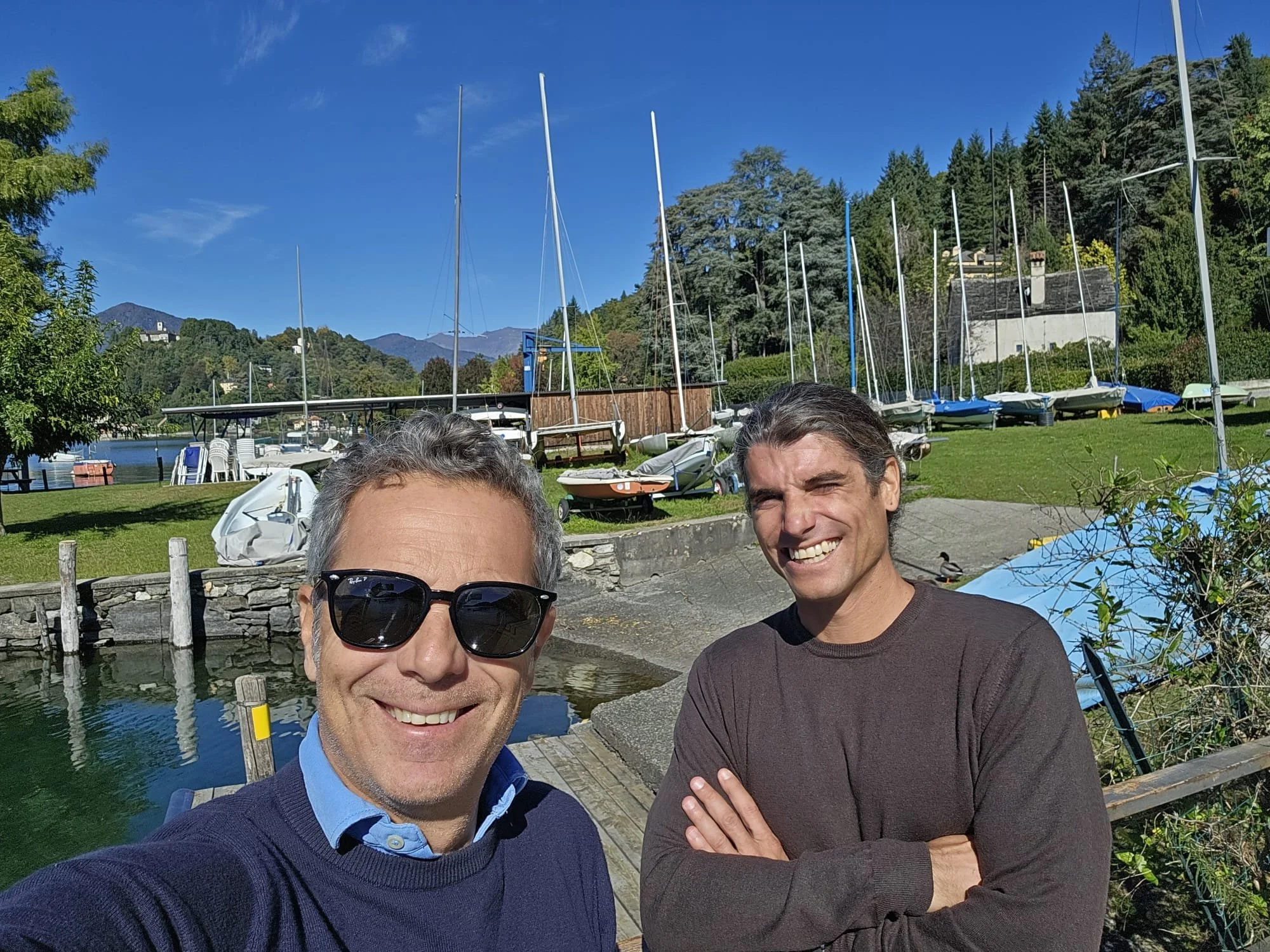 Due uomini sorridenti si trovano vicino a un lago con barche ormeggiate, in un ambiente naturale con alberi e montagne sullo sfondo in una giornata di sole.
