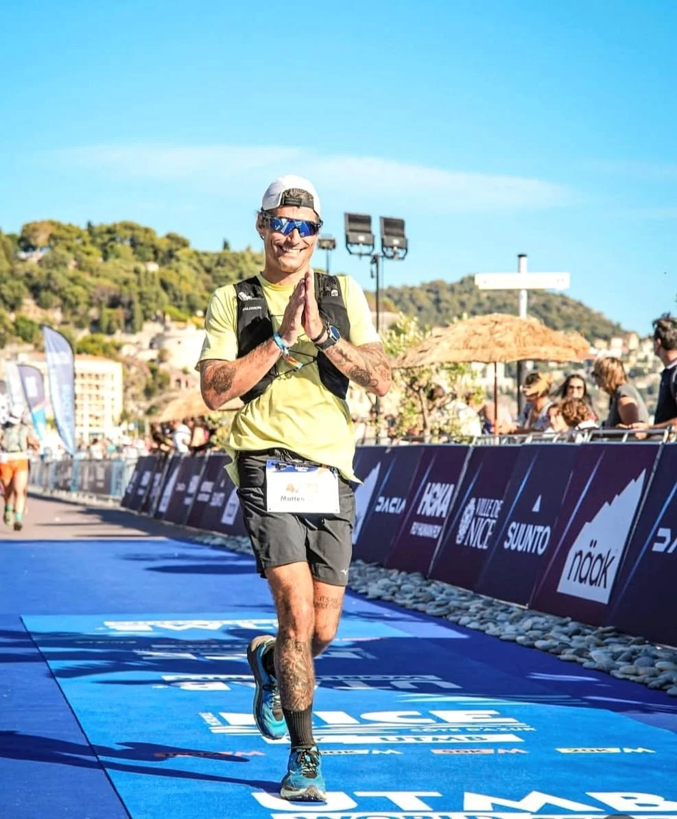 Accompagnatore e  bike che corre e sorride con le mani giunte in segno di preghiera o di gratitudine durante una maratona, con pubblico ai lati e un paesaggio di colline verdi sullo sfondo.