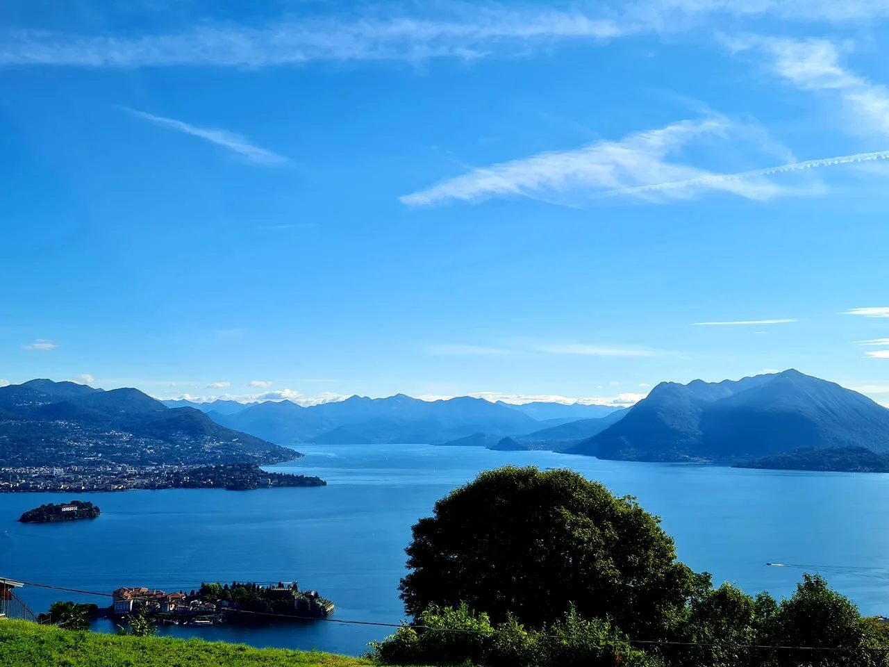Paesaggio di uno splendido lago circondato da montagne con un cielo sereno. In primo piano ci sono alberi e una zona verde.