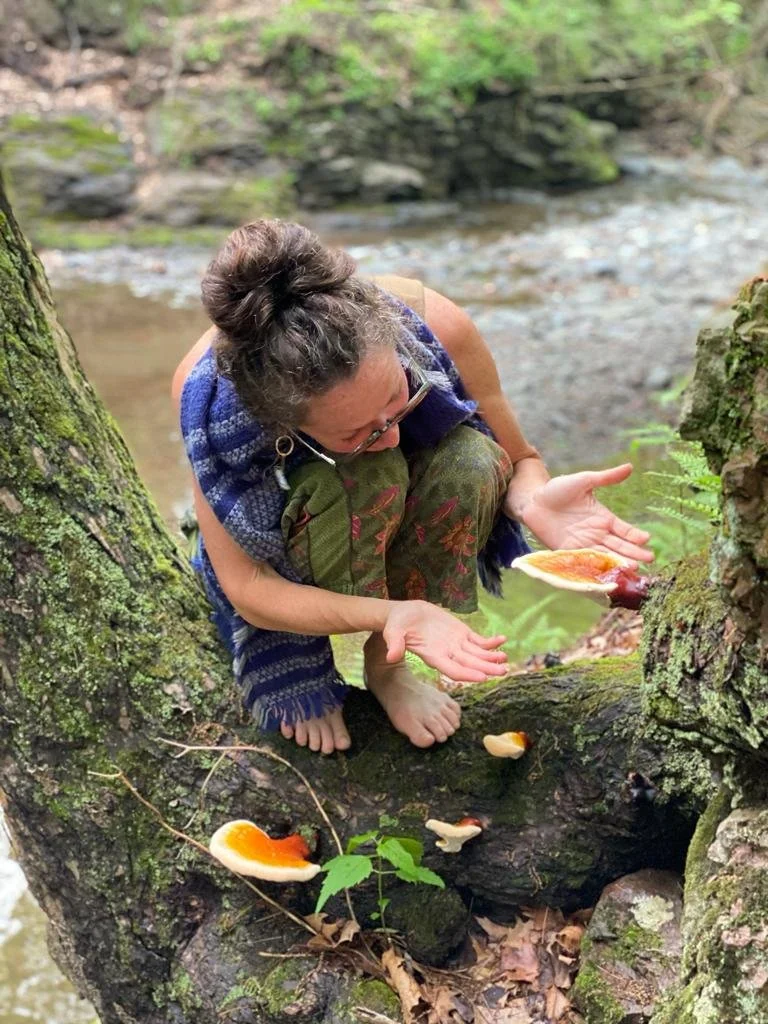 A woman with glasses and curly hair, wearing a blue scarf and camouflage pants, crouches on a mossy tree branch by a creek, holding a mushroom in her open hands.