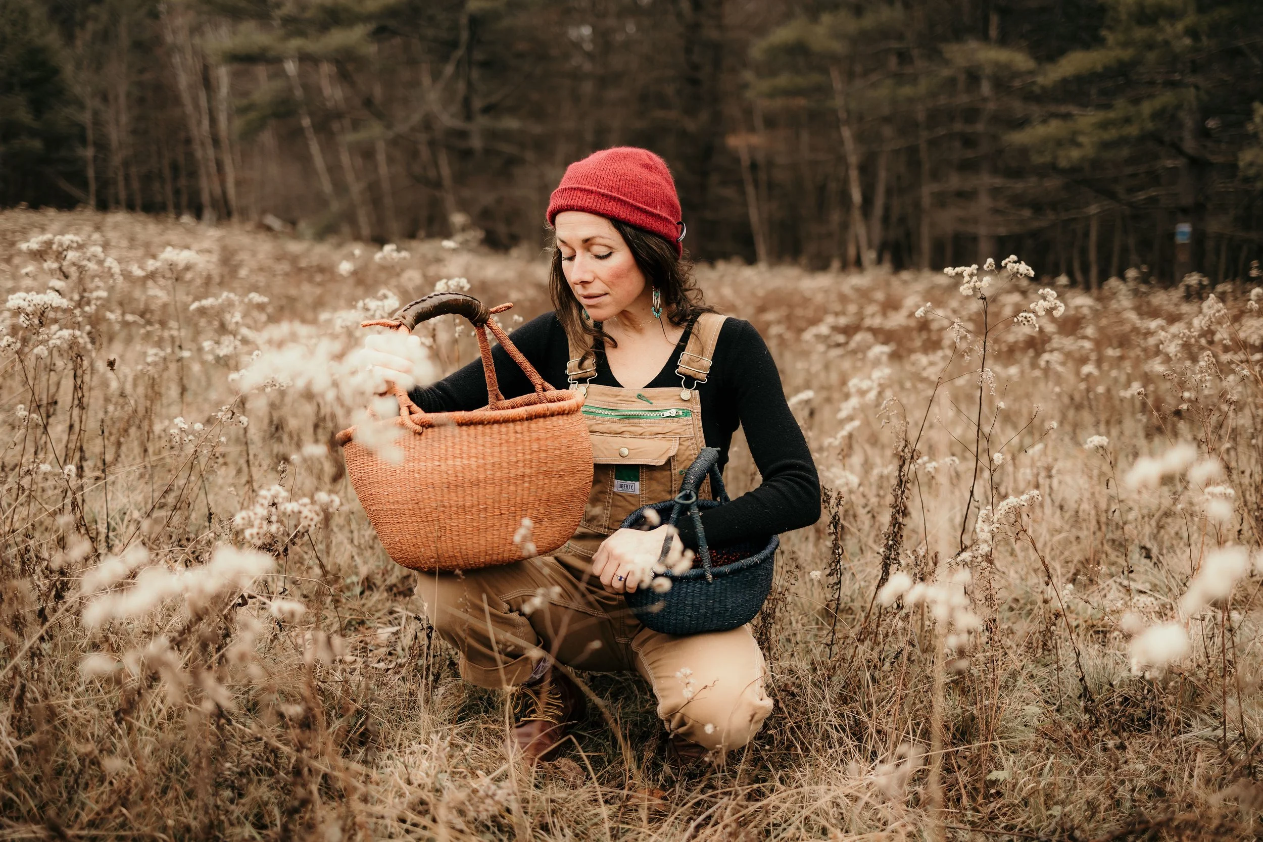 A woman wearing a red beanie, black long-sleeve shirt, and brown overalls, crouching in a field of dry grass and white wildflowers with a forest in the background. She is holding baskets while examining the flowers.
