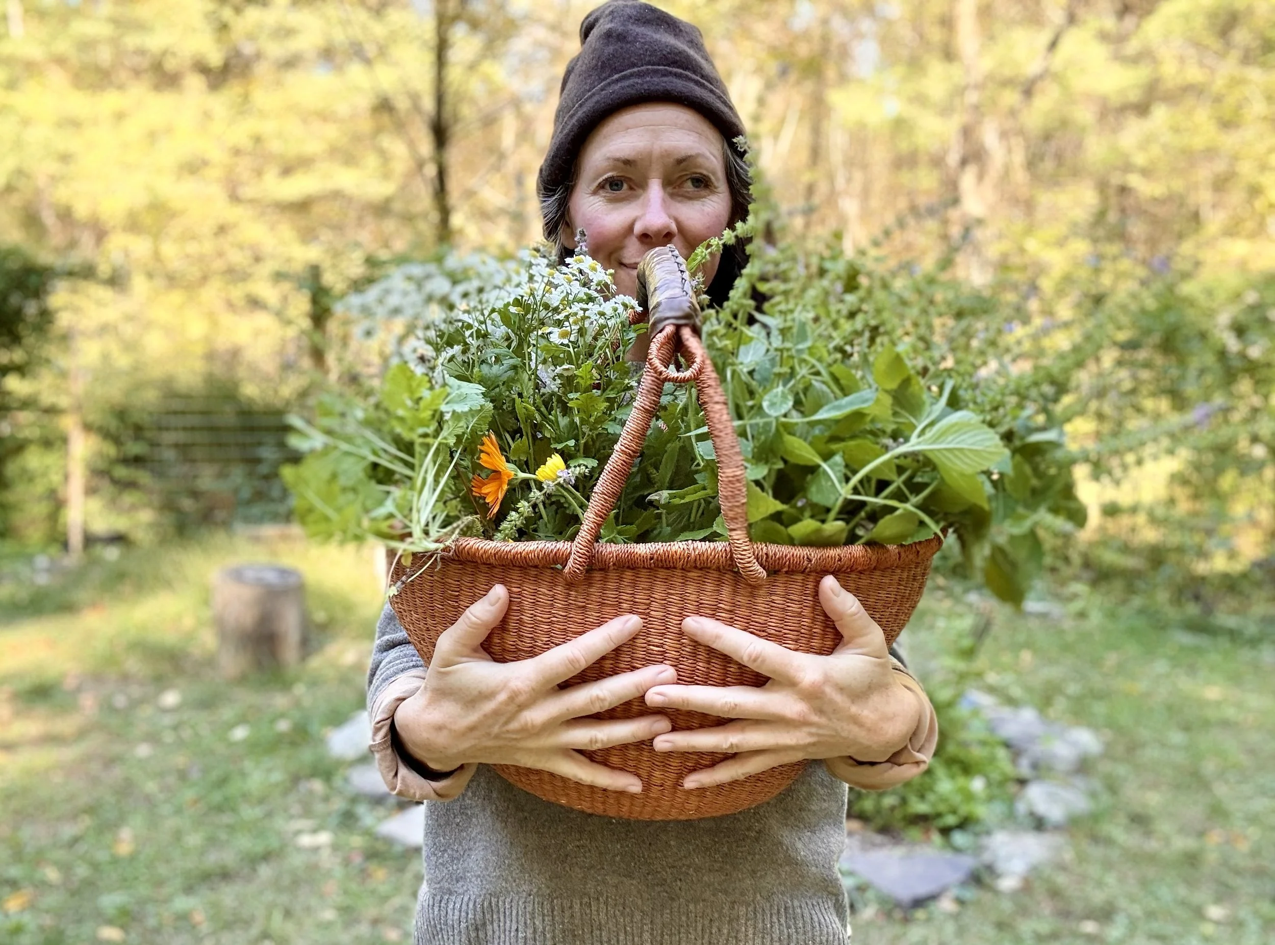 A woman wearing a black beanie and gray sweater holding a large wicker basket filled with green plants and flowers outdoors in a garden with trees in the background.