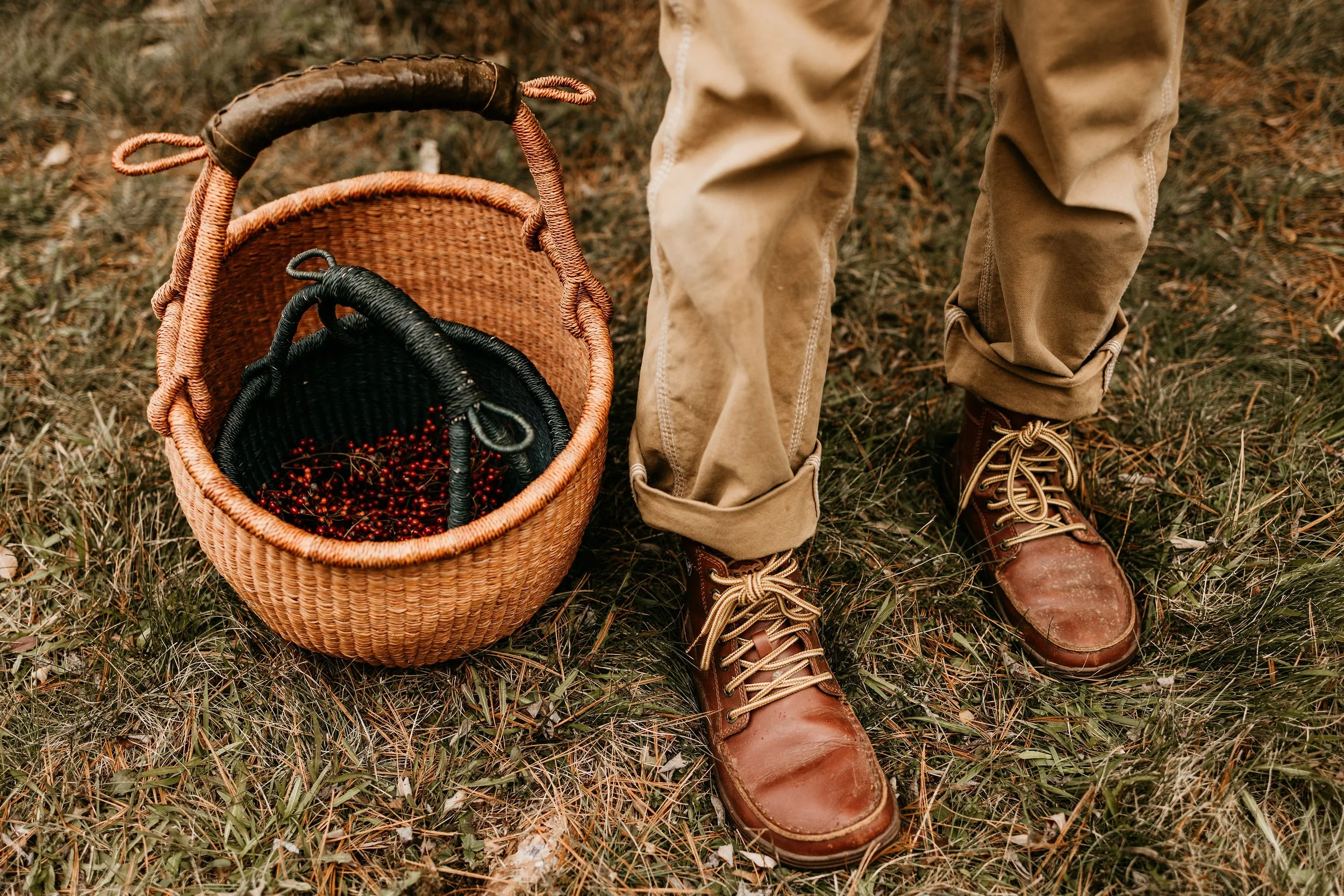 Person standing outdoors on grass with a woven basket filled with red berries, wearing tan pants and brown lace-up boots.