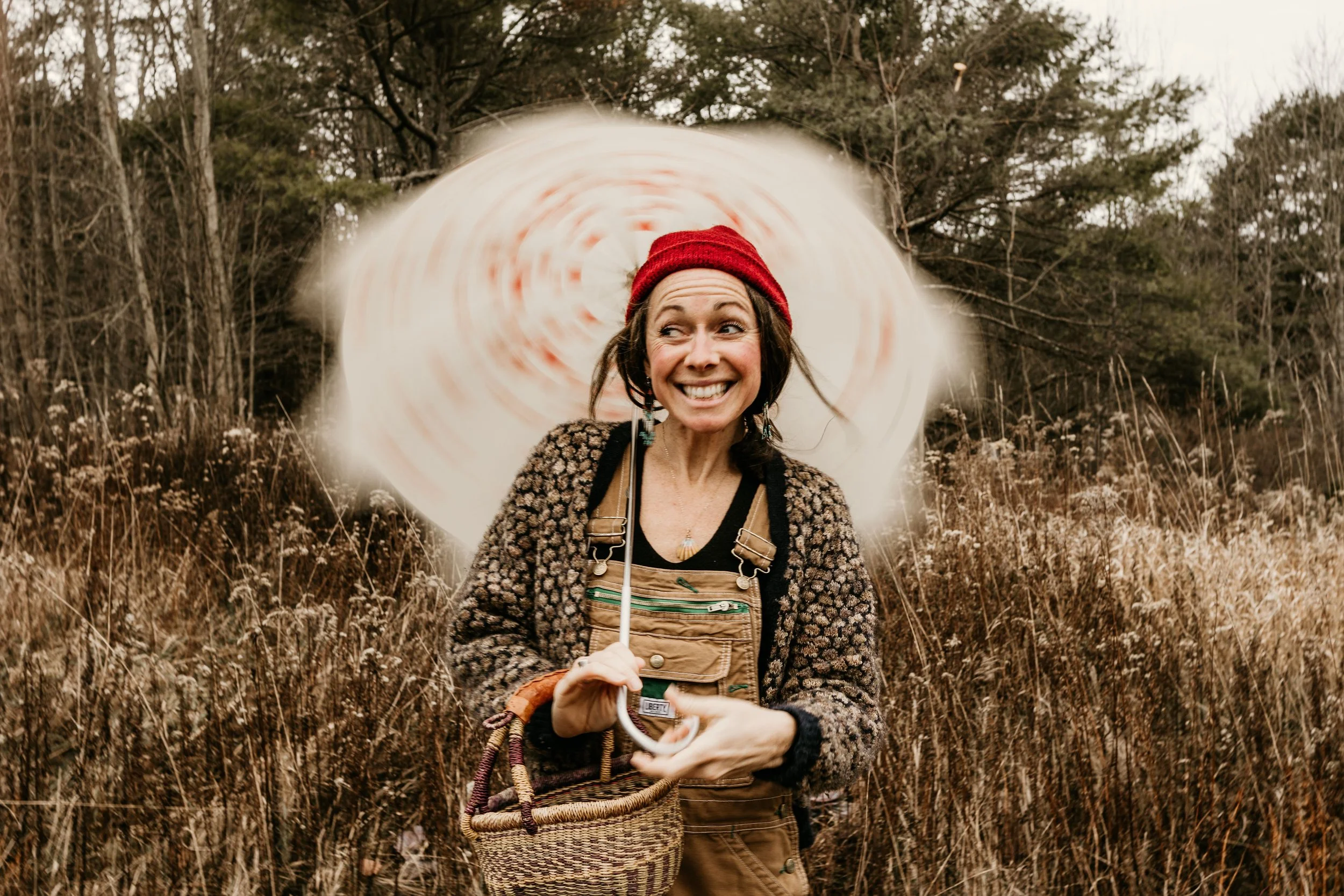 A woman smiling and holding an umbrella outdoors in a field with tall grasses and trees in the background.