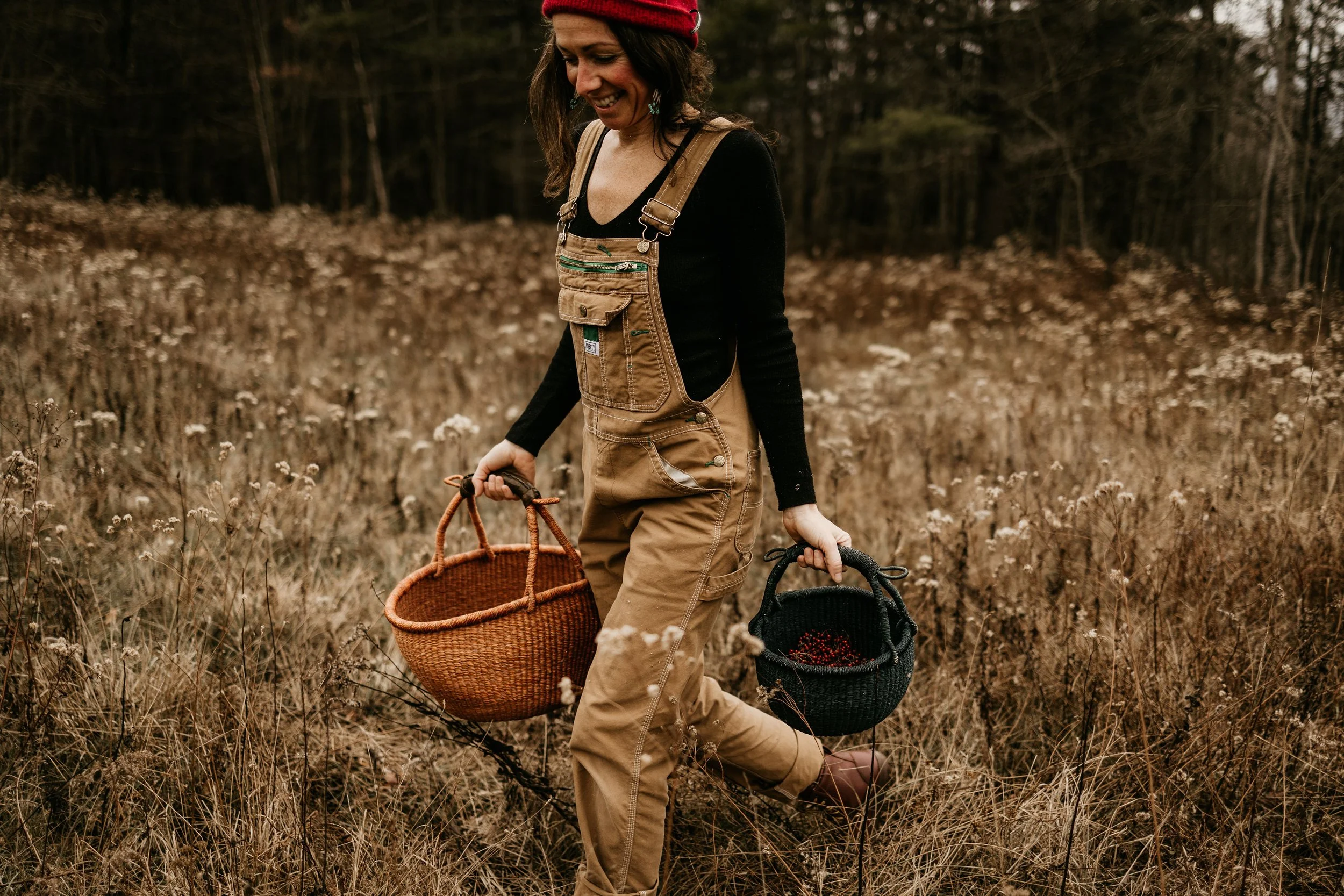 A woman walking through a field with two baskets, one in each hand, wearing overalls and a red beanie, smiling and looking down.