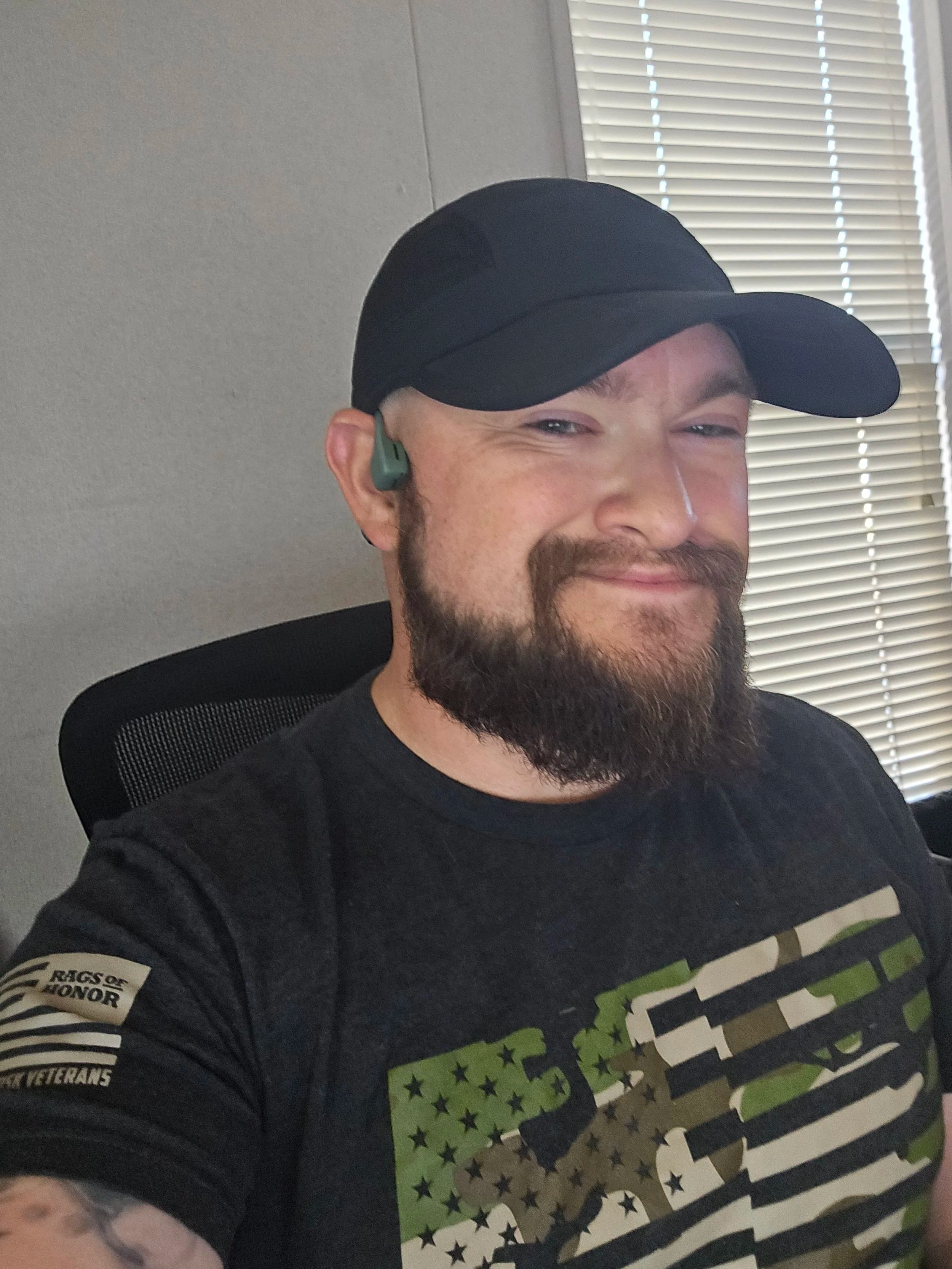A smiling man with a beard wearing a black cap, a black t-shirt with a camouflage American flag design, and a green and black Bluetooth earpiece, sitting in an office chair near a window with blinds.
