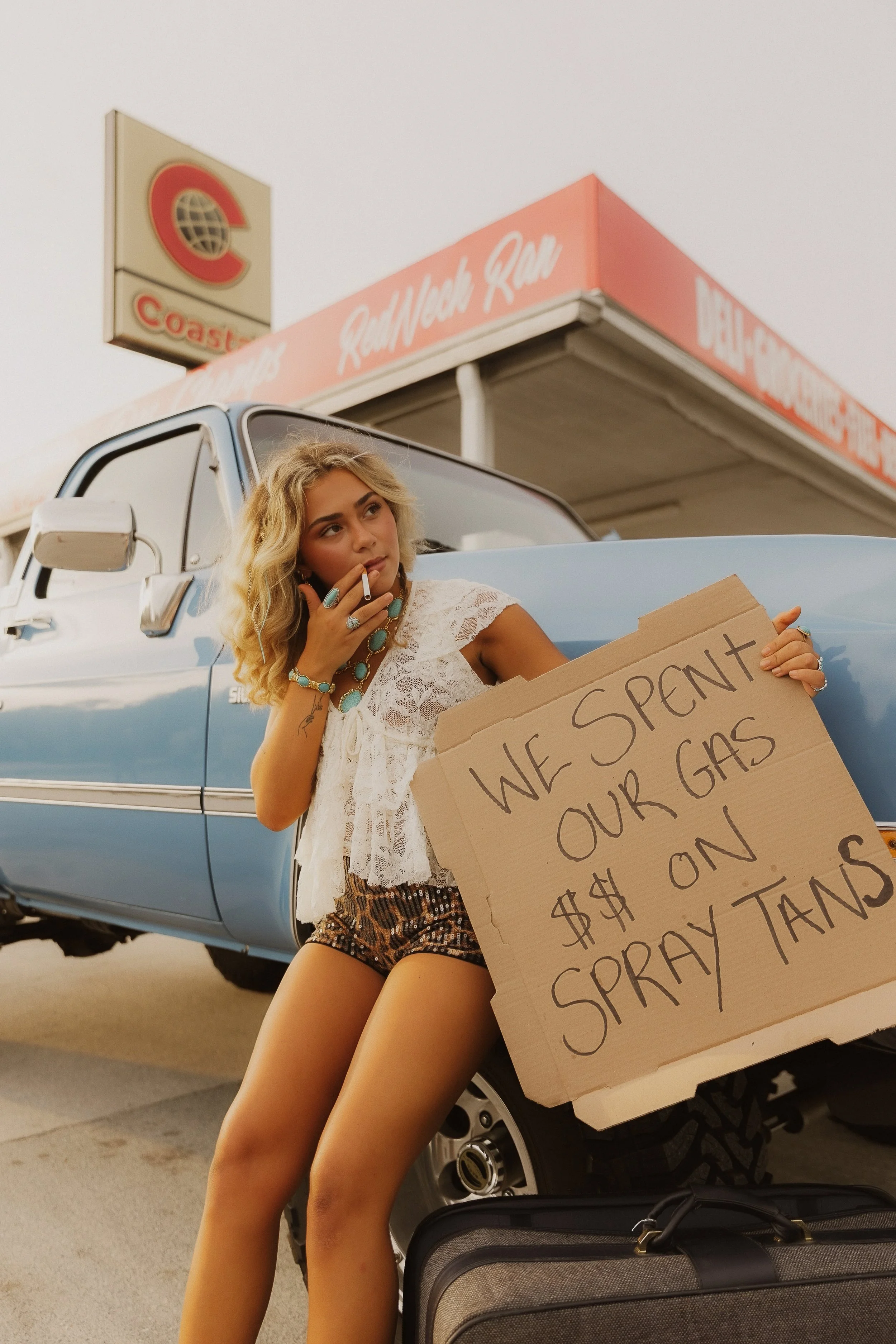 A young woman with curly blonde hair, wearing jewelry and a white lace top, sits on the ground next to a blue pickup truck. She holds a cardboard sign with handwritten text about gasoline and money. She is smoking a cigarette while looking thoughtfully away. There is a suitcase on the ground beside her and a gas station sign in the background.