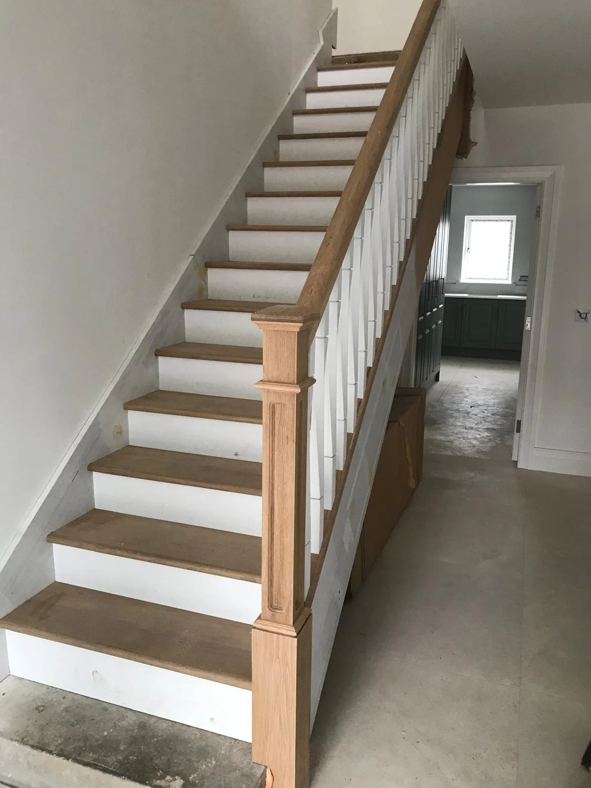 Incomplete wooden staircase in a house, with some steps painted white and some unfinished, leading to an upper floor. The staircase has a wooden handrail and newly installed balusters. The background shows a doorway to a room with green cabinetry, a