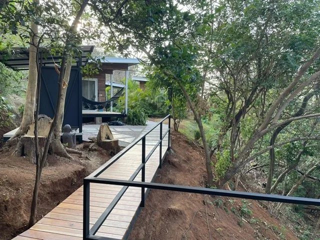 Wooden walkway with black metal railings on a hillside surrounded by trees and vegetation, leading to a modern house with outdoor seating and a hammock.