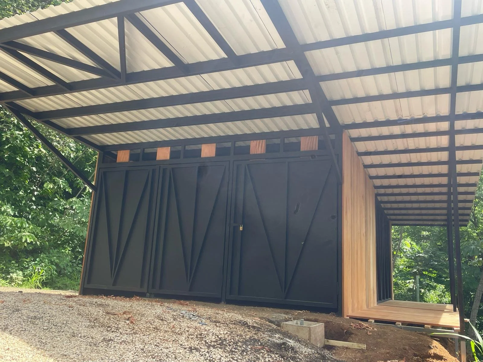 A modern outdoor structure with a black metal frame, wooden walls, and a metal roof, surrounded by green trees.