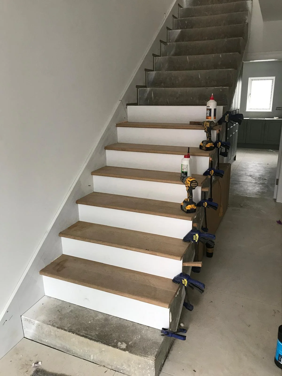 Under construction staircase with wooden treads and white risers, clamps holding the steps, construction tools and supplies on the side, and unfinished concrete floor.