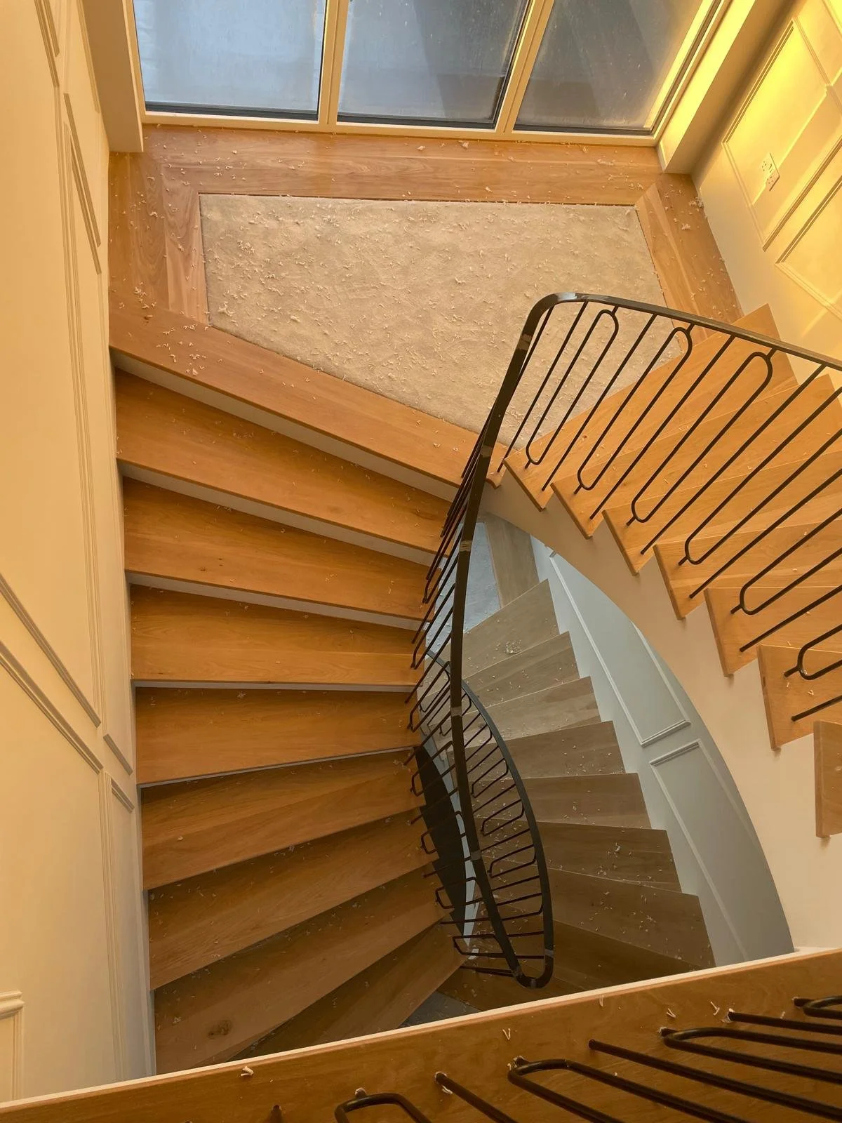 Overhead view of a spiral staircase with wooden steps and black railing, overlooking a small carpeted landing at the bottom, inside a house with a sliding glass door.
