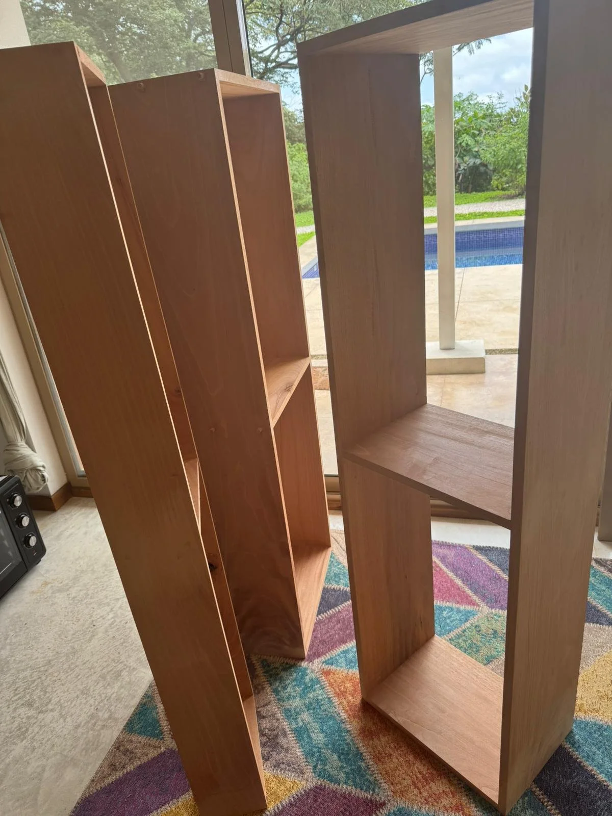 Two wooden bookshelves next to each other inside a room, with a sliding glass door and a backyard with a pool outside.