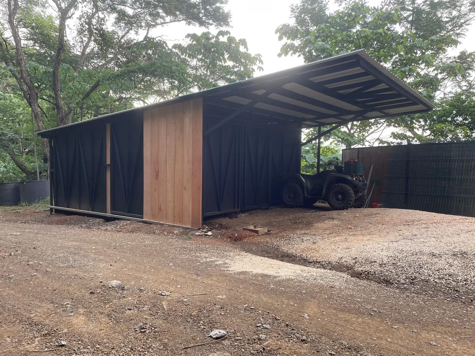 A partially enclosed shed with black metal sides, a roof, and a wooden panel in a yard with gravel ground, surrounded by trees. There is a four-wheeler parked inside the shed.