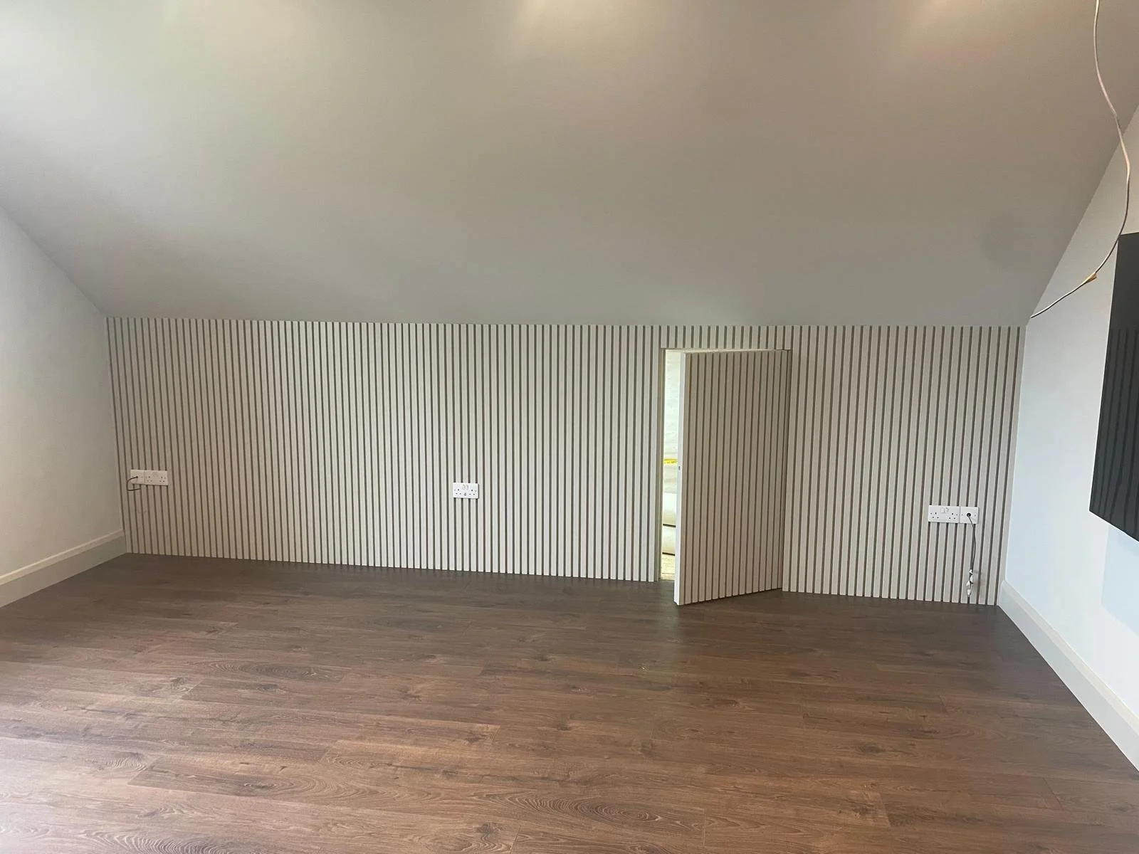 An empty room with a sloped ceiling, striped wallpaper on one wall, three electrical outlets, and a small doorway leading to a balcony or patio.