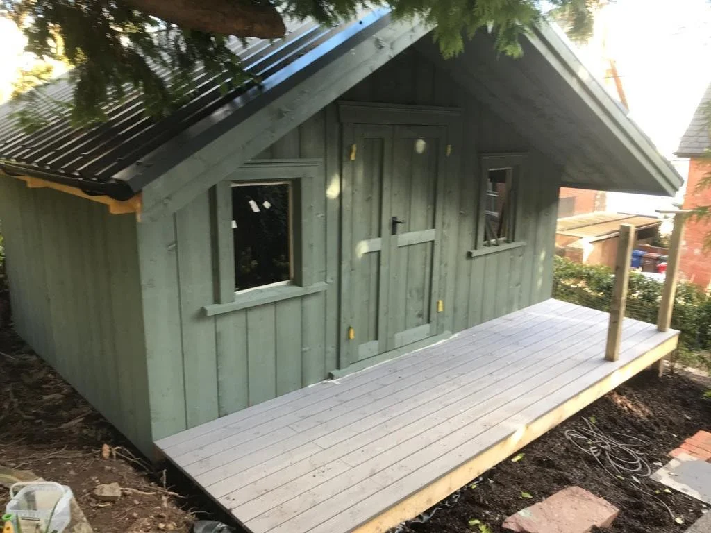 Small wooden shed with green exterior, two small windows, and a door, with a new deck in front, surrounded by trees and neighboring houses.