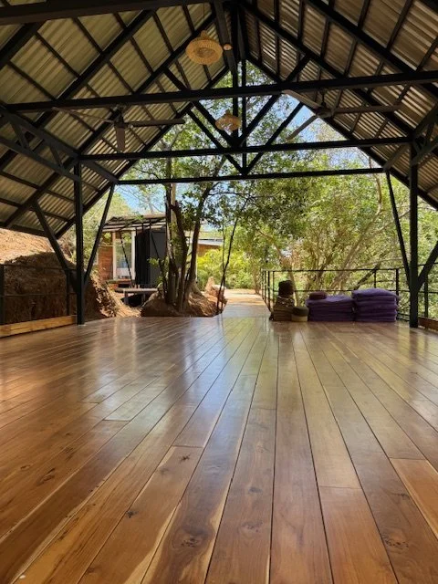 Interior of a covered outdoor space with wooden flooring, metal framework, hanging lights, and view of trees and a small structure outside.